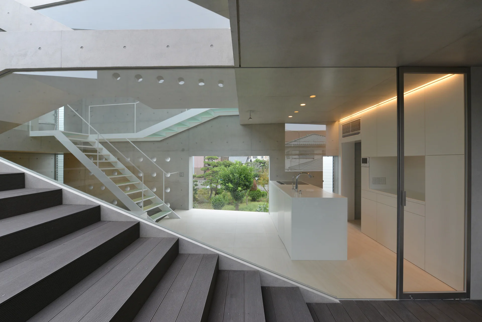 A modern minimalist kitchen features a white island and cabinetry set against raw concrete walls, framed by a dark wood staircase and glass divider.
