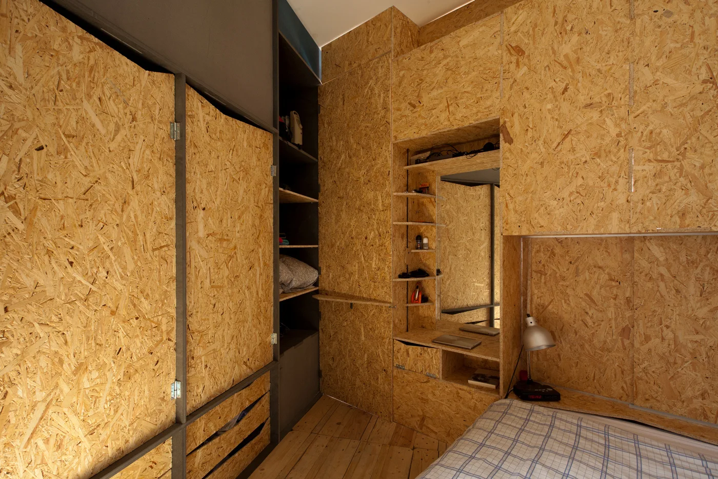 A compact bedroom features extensive custom built-in storage cabinets made from oriented strand board, alongside a small bed and vanity mirror niche.
