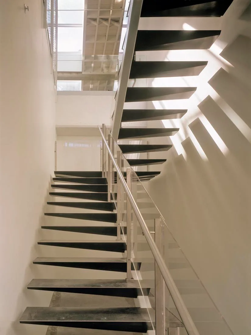 A modern black metal floating staircase with a glass railing winds upward in a white stairwell under a skylight casting geometric shadows on the wall.