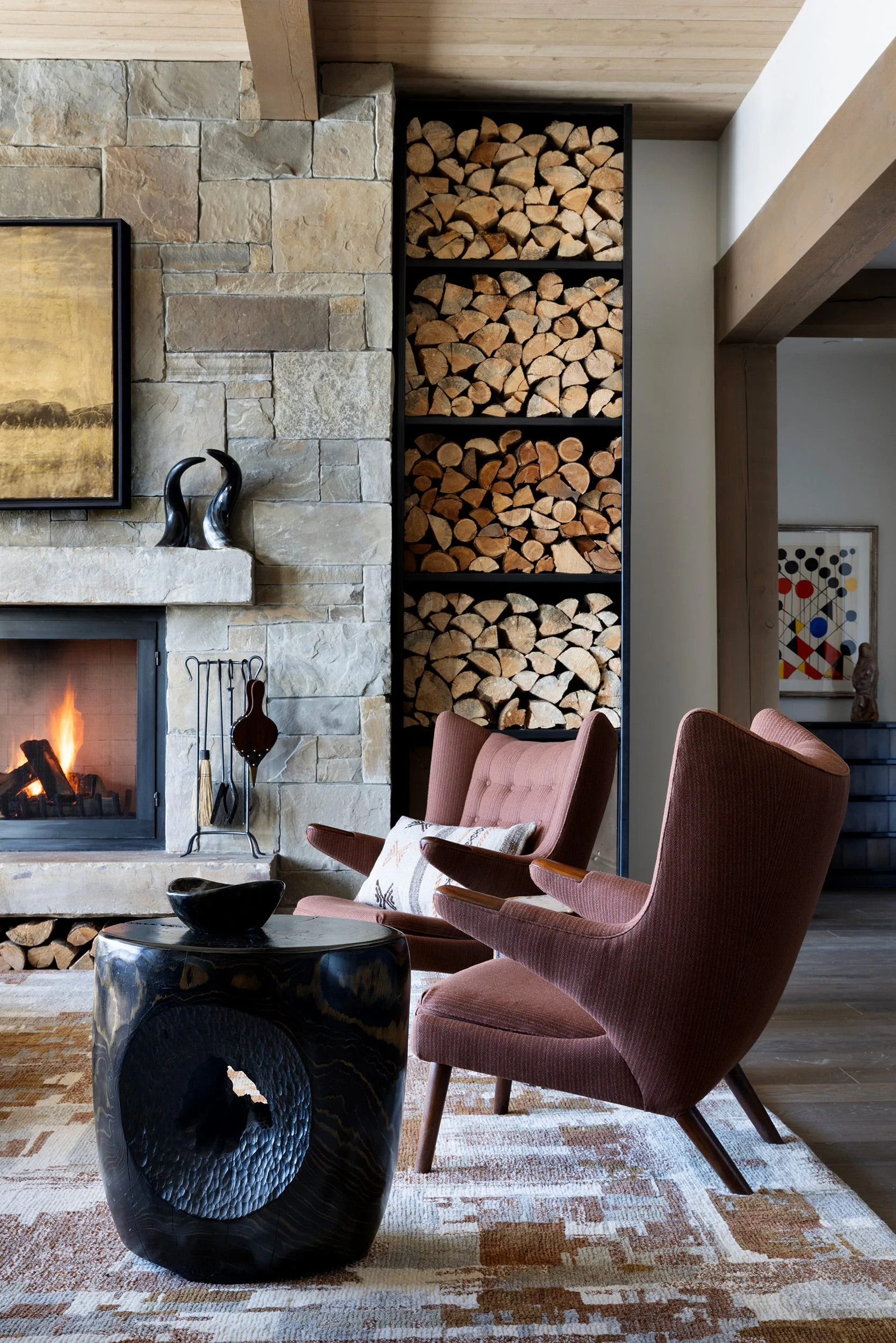 A cozy living room features a stone fireplace next to a tall black niche stacked with firewood and two brown wingback armchairs.