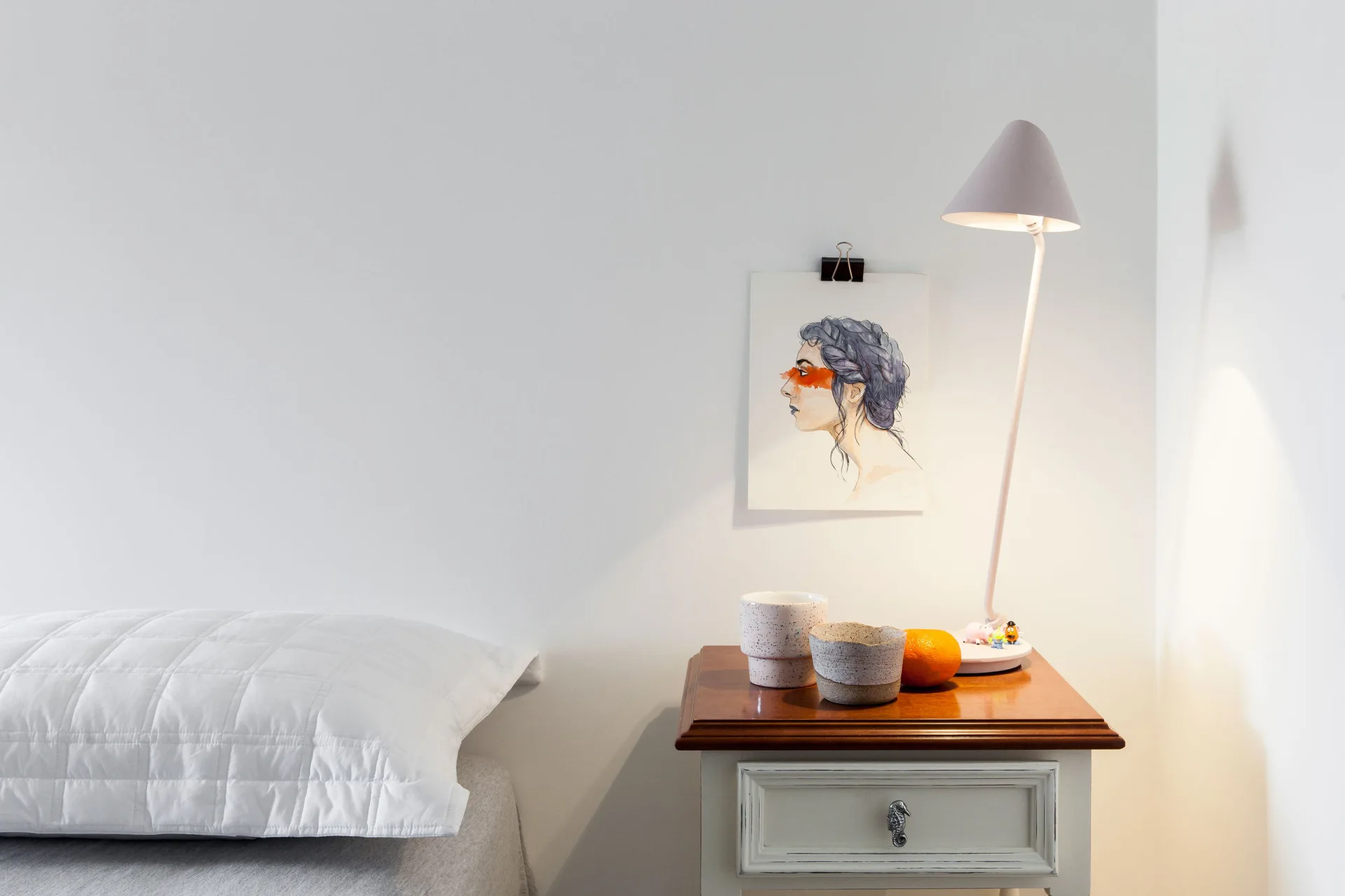 A white bedside nightstand with a wood top holds a slim table lamp, speckled ceramic bowls, and an orange next to a quilted white pillow.