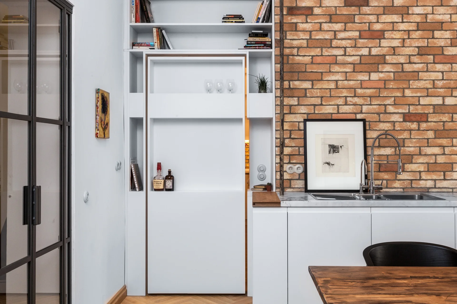 A modern kitchen features white cabinets, an exposed brick wall with a sink, and a tall white custom shelving unit with a sliding door panel.