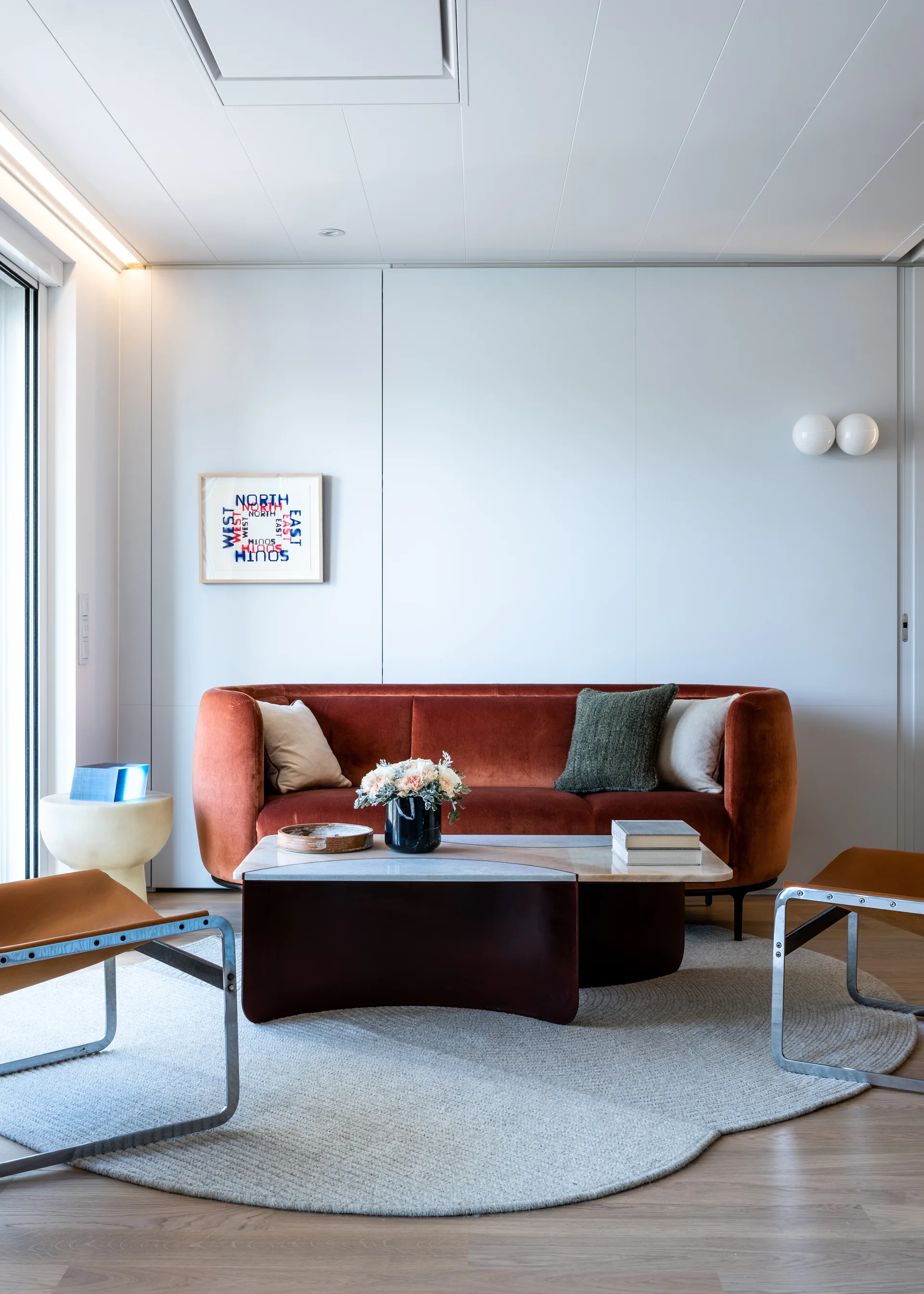 A rust velvet sofa stands on a round woven rug opposite two leather armchairs and a modern coffee table against a smooth white wall.