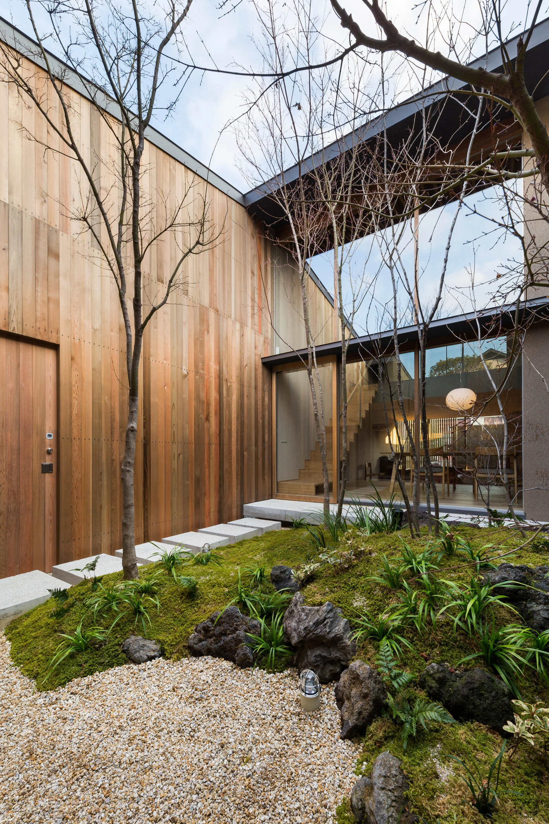 A modern residential courtyard features a mossy rock garden, stone stepping stones, and a vertical wood-paneled wall next to a large glass window.