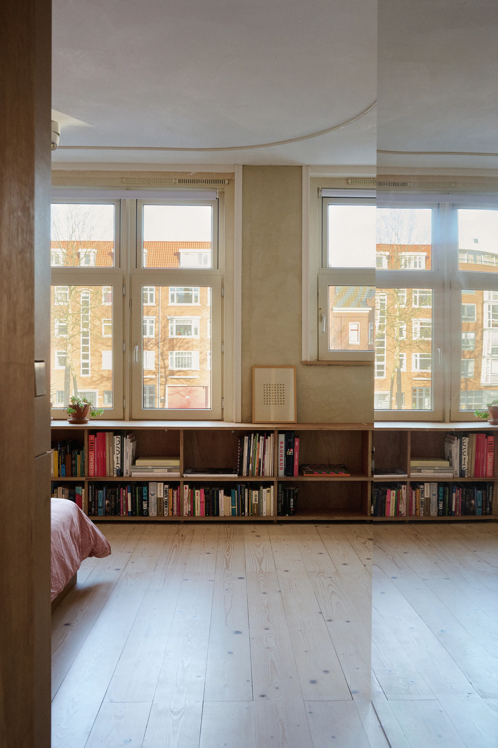 A minimalist bedroom features low wooden built-in bookshelves positioned under large windows, alongside light wood plank flooring.