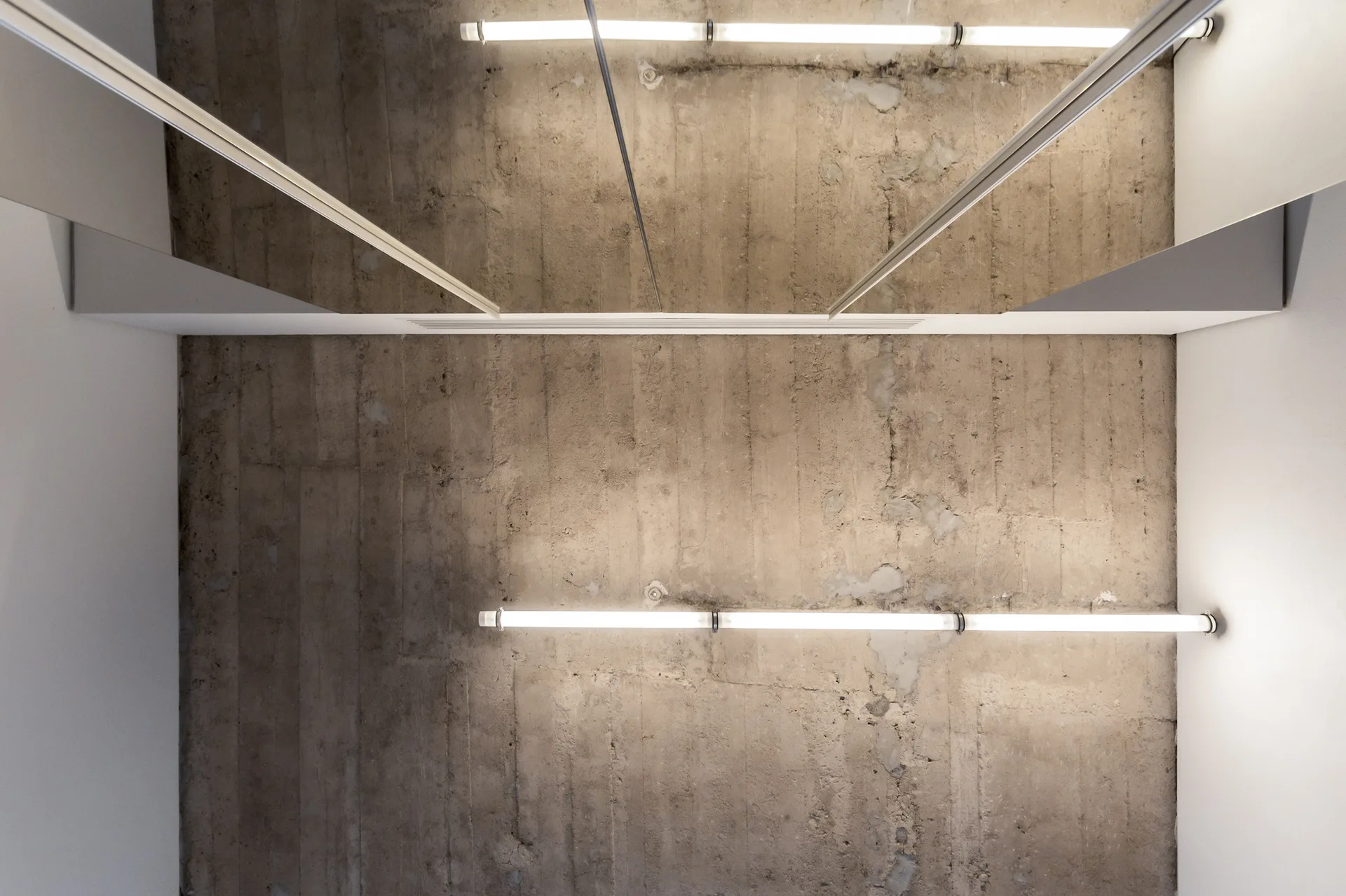 A low-angle view of a textured concrete ceiling with two mounted linear light fixtures positioned between smooth white walls and reflective mirrors.