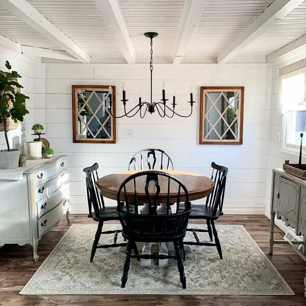A round wooden dining table surrounded by black spindle chairs sits on a patterned rug under a black chandelier in a white shiplap dining room.