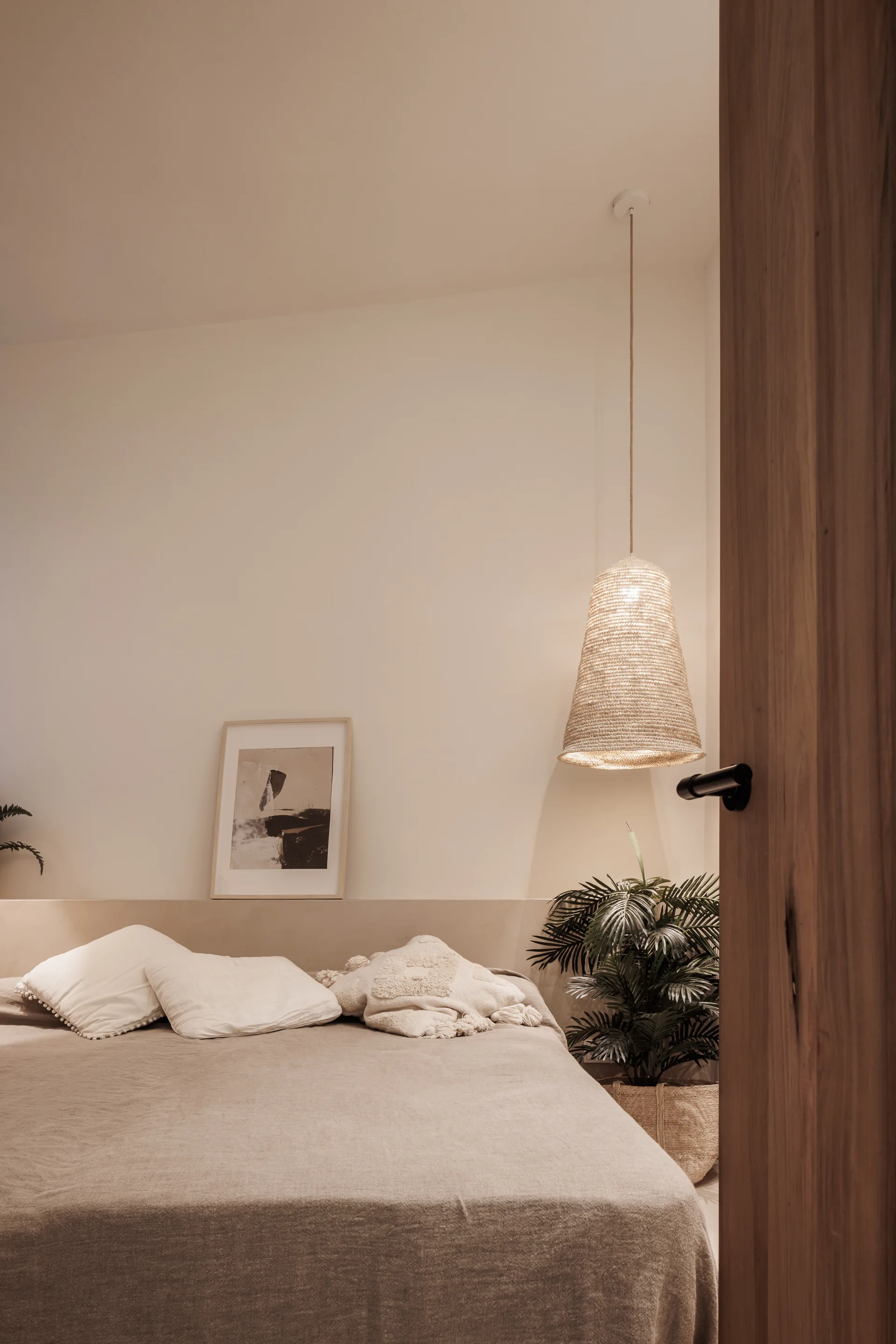 A neutral bedroom viewed through a wooden doorway, featuring a bed with white textured pillows, a low ledge headboard, and a hanging woven pendant light.