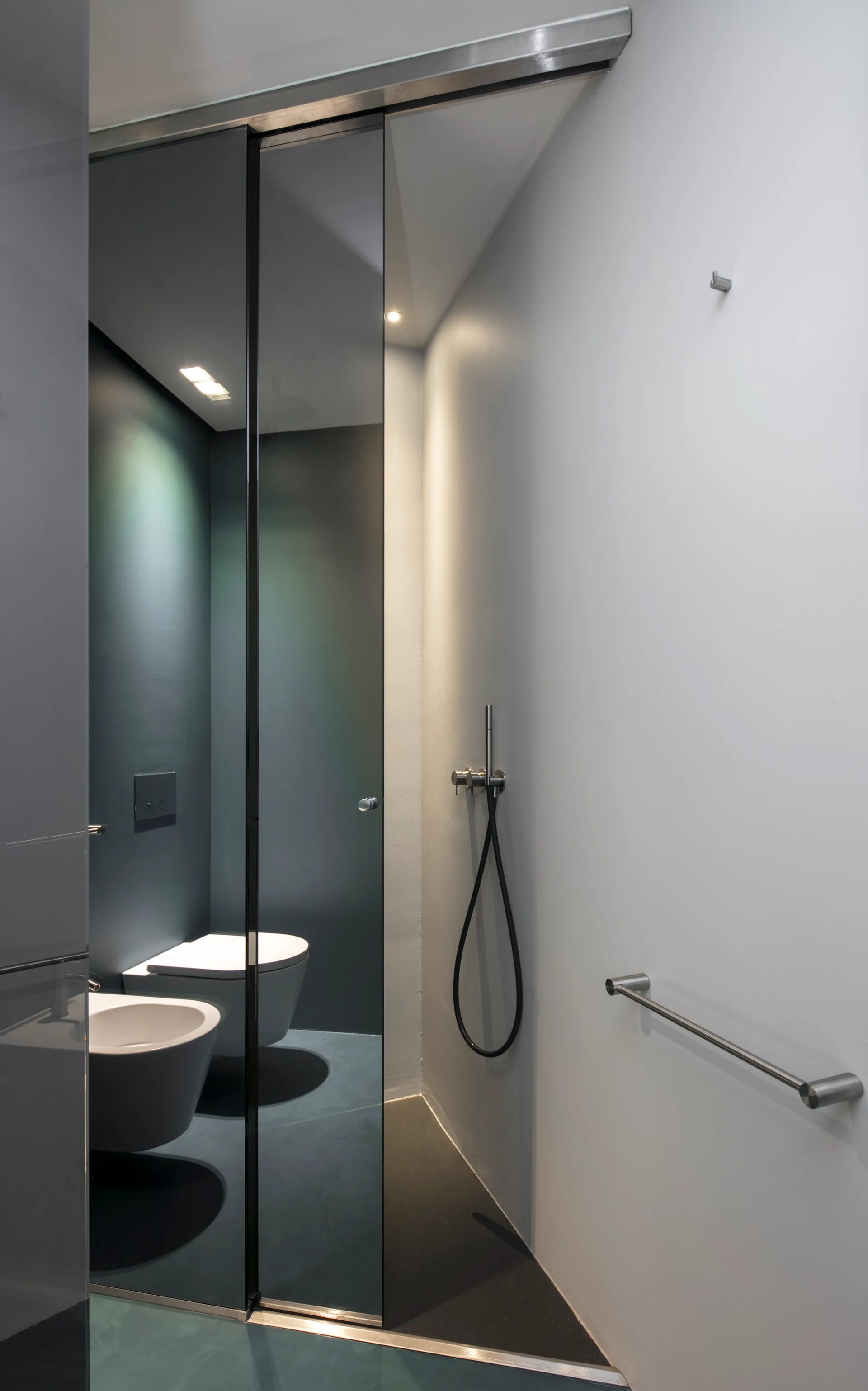 A minimalist modern bathroom features a dark-tinted glass sliding partition next to a wall-mounted toilet and bidet.