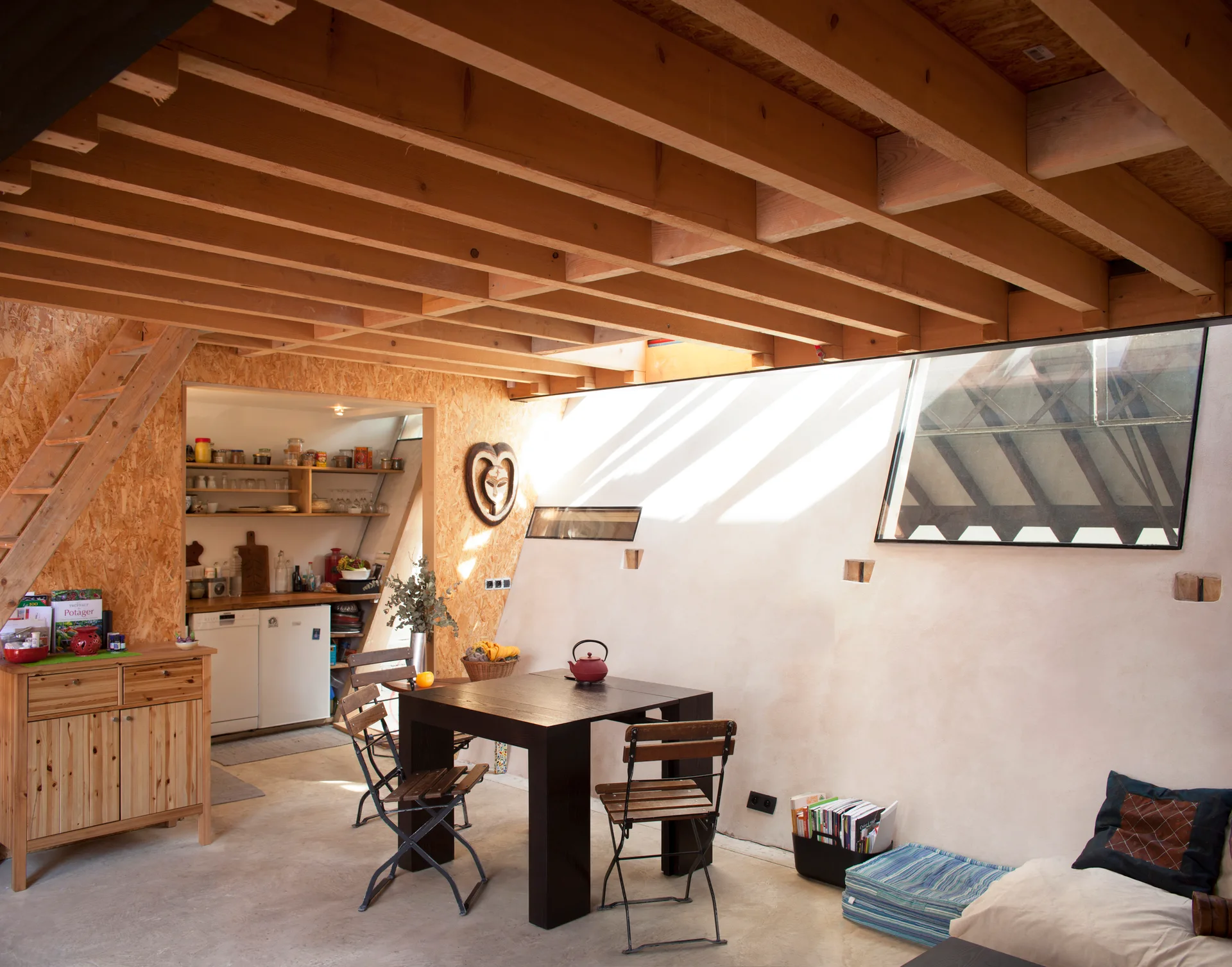 A dining area with a dark wood table beneath exposed timber rafters, featuring OSB-paneled walls and a sloped skylight.