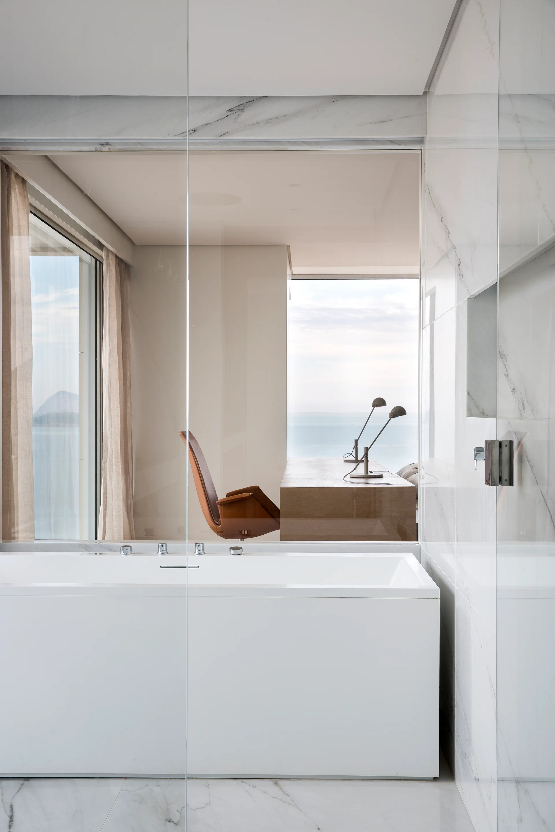 A white marble bathroom with a square bathtub is separated by a clear glass wall from a home office with a wood desk overlooking the ocean.