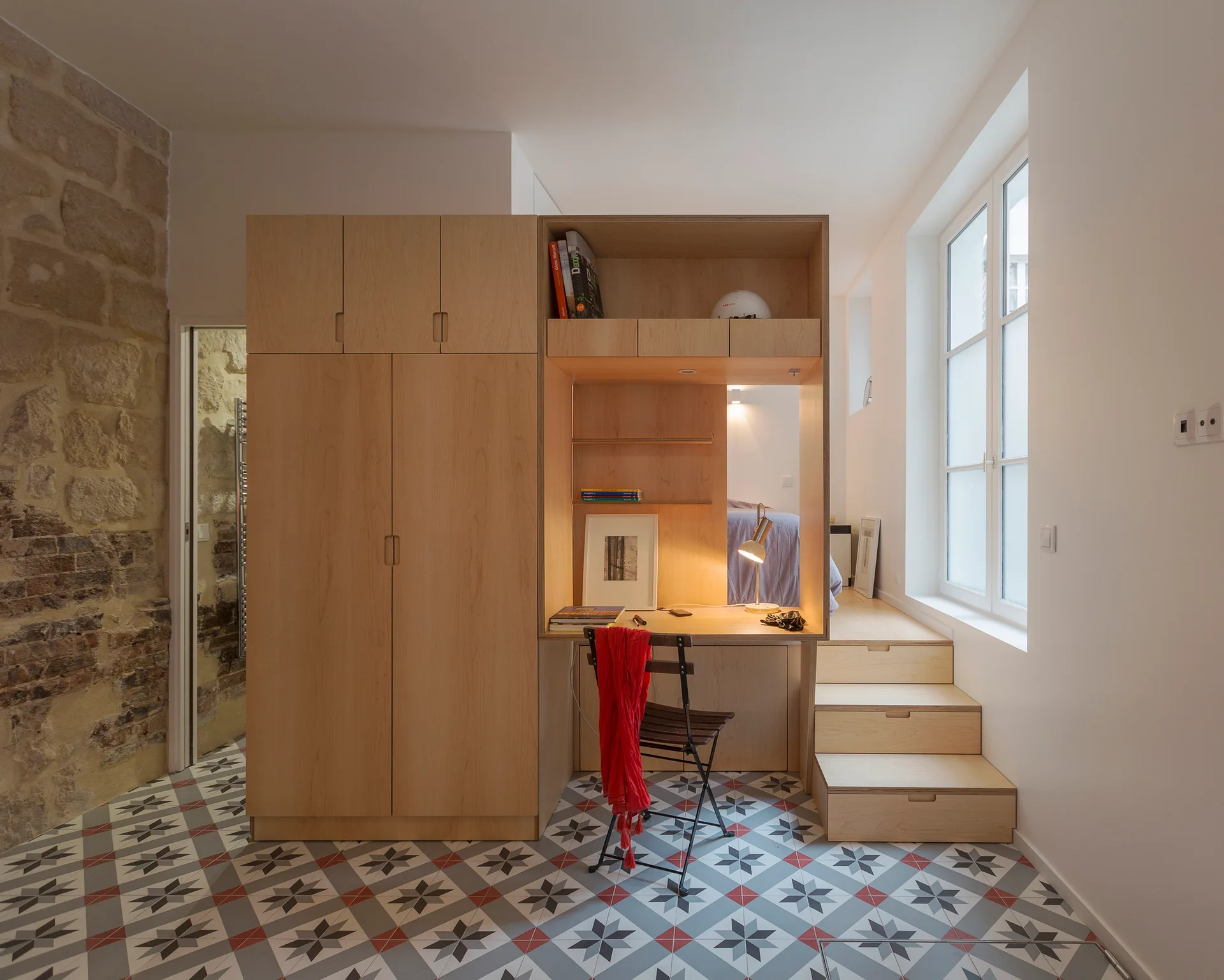 A custom light wood wardrobe and desk unit stands next to a wooden stepped platform leading to a raised bed in a room with patterned tile floors.