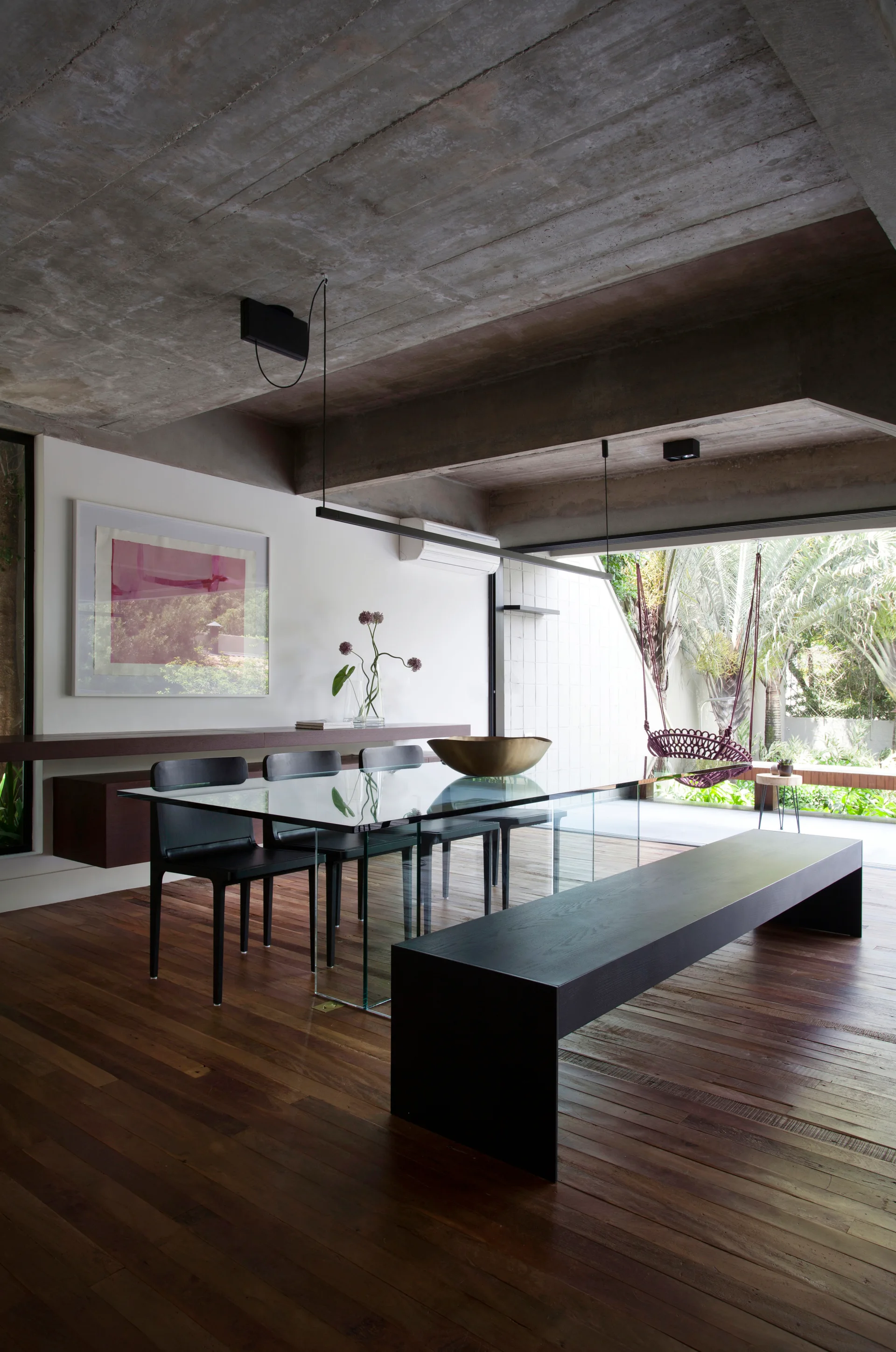 A minimalist dining room features a glass-topped dining table, black chairs, a dark wood bench, and an exposed raw concrete ceiling.