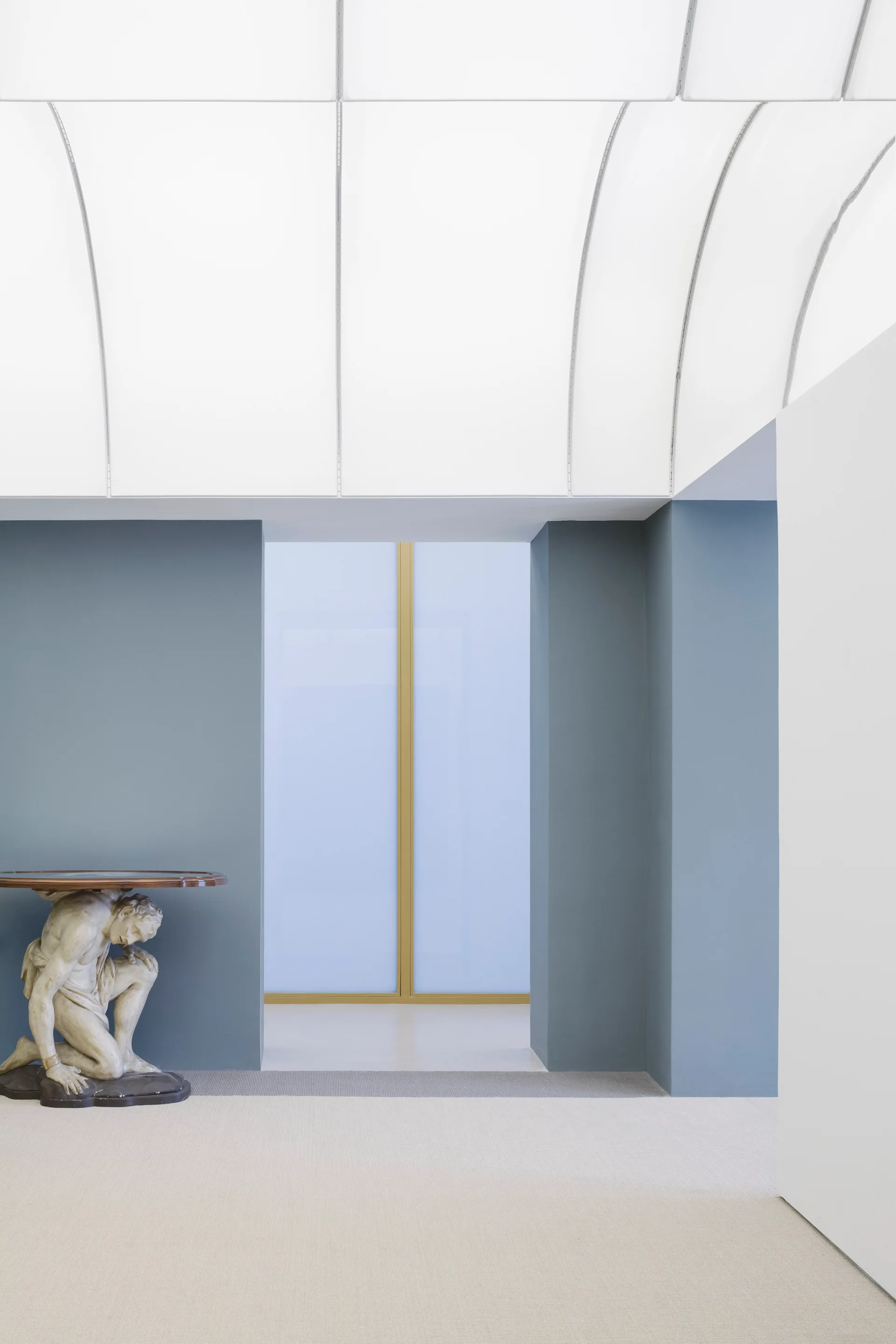 A minimalist hallway features blue walls, a curved white ceiling, and a console table with a classical figurative sculpture base.