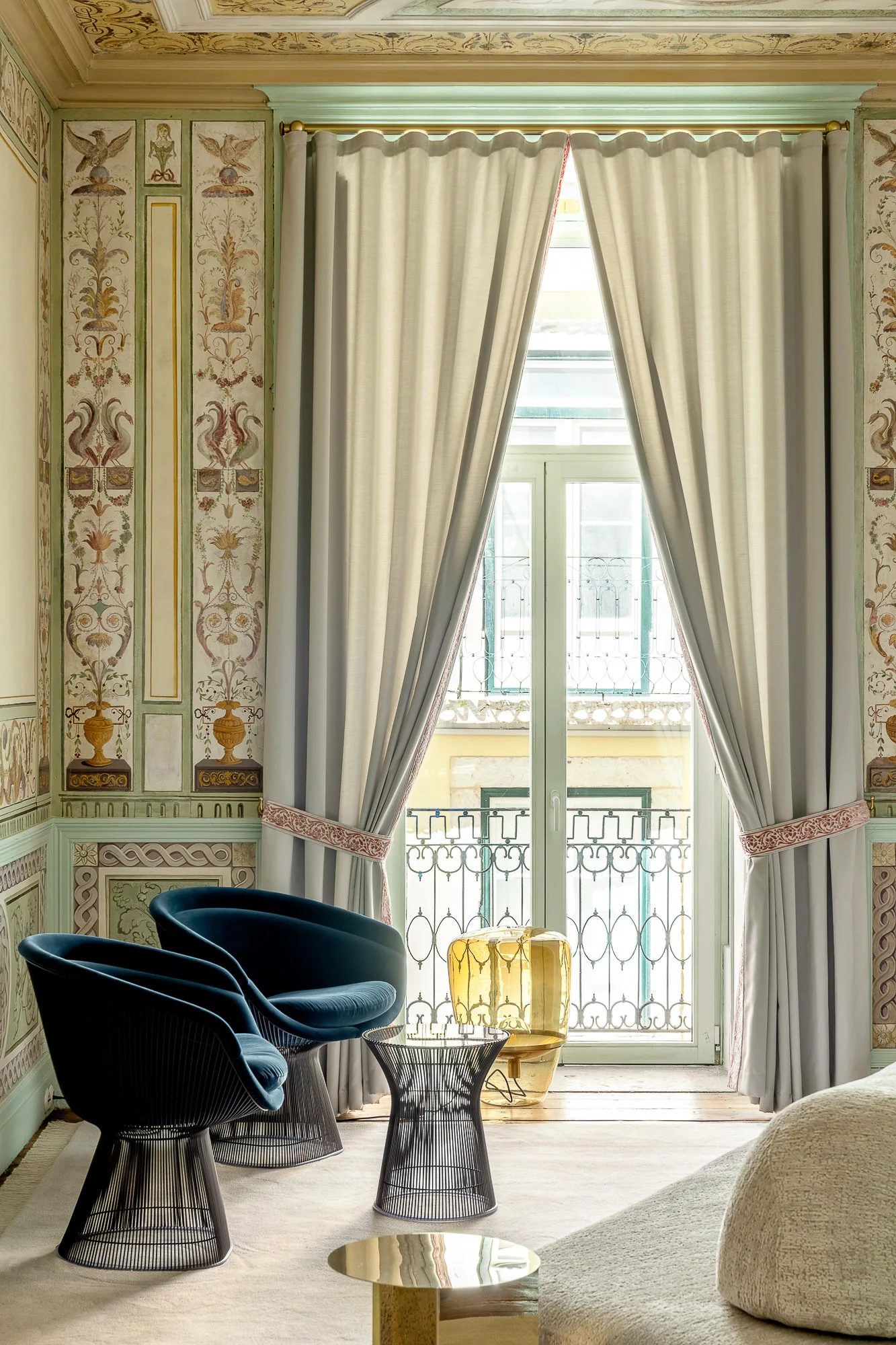 A bright sitting area in a historic room features ornate painted wall panels, two deep blue velvet armchairs, and floor-length beige curtains.
