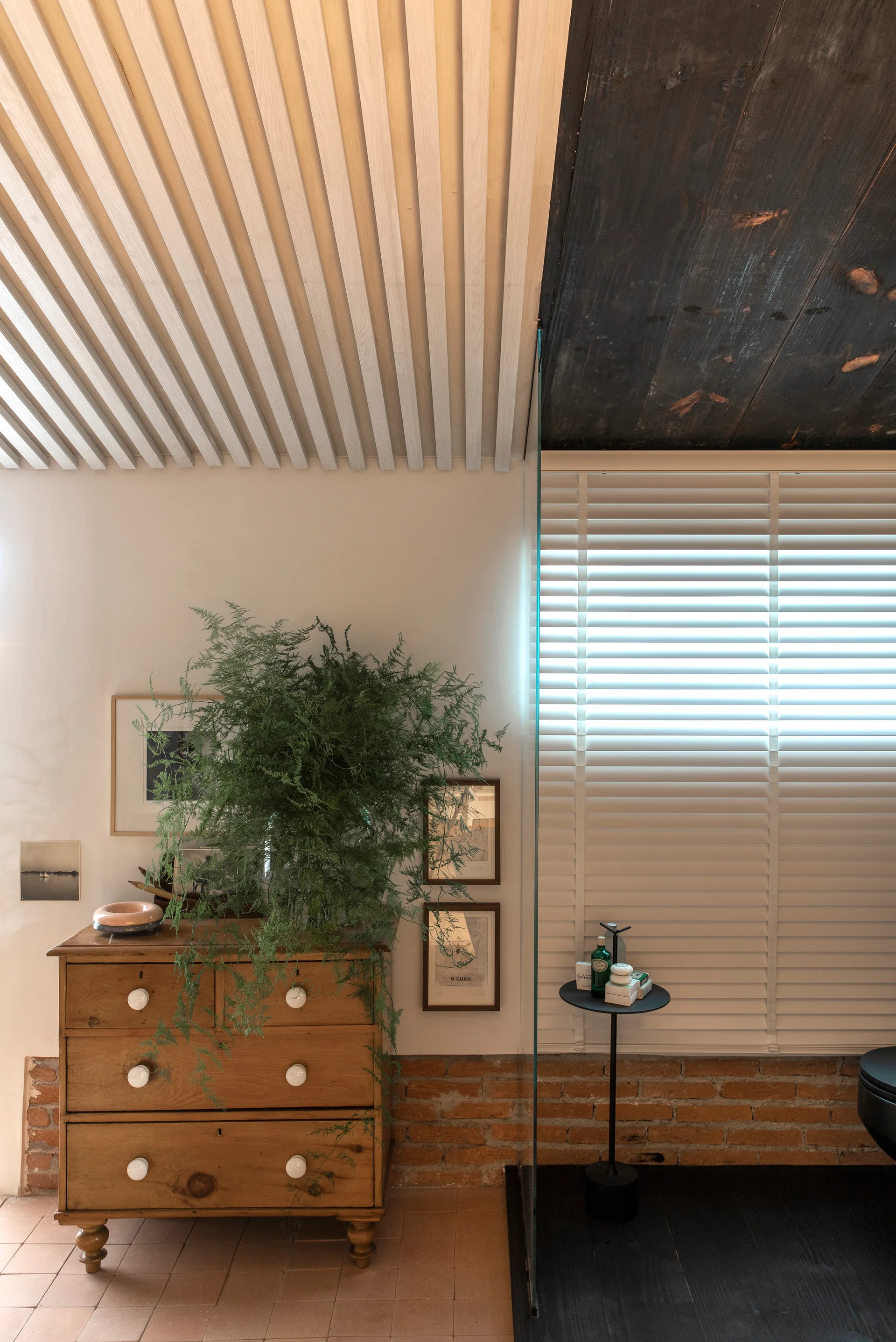 A vintage wooden dresser with a leafy green plant sits against a white plaster wall with a base of exposed red brick.