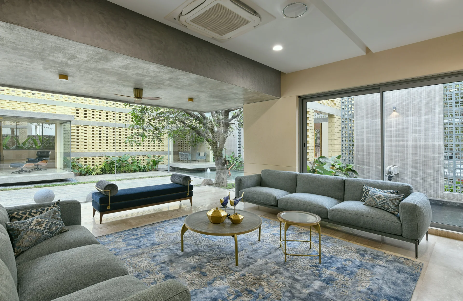 A modern living room featuring two gray sofas, a dark blue bolster bench, gold coffee tables, and a large patterned rug next to floor-to-ceiling glass walls.