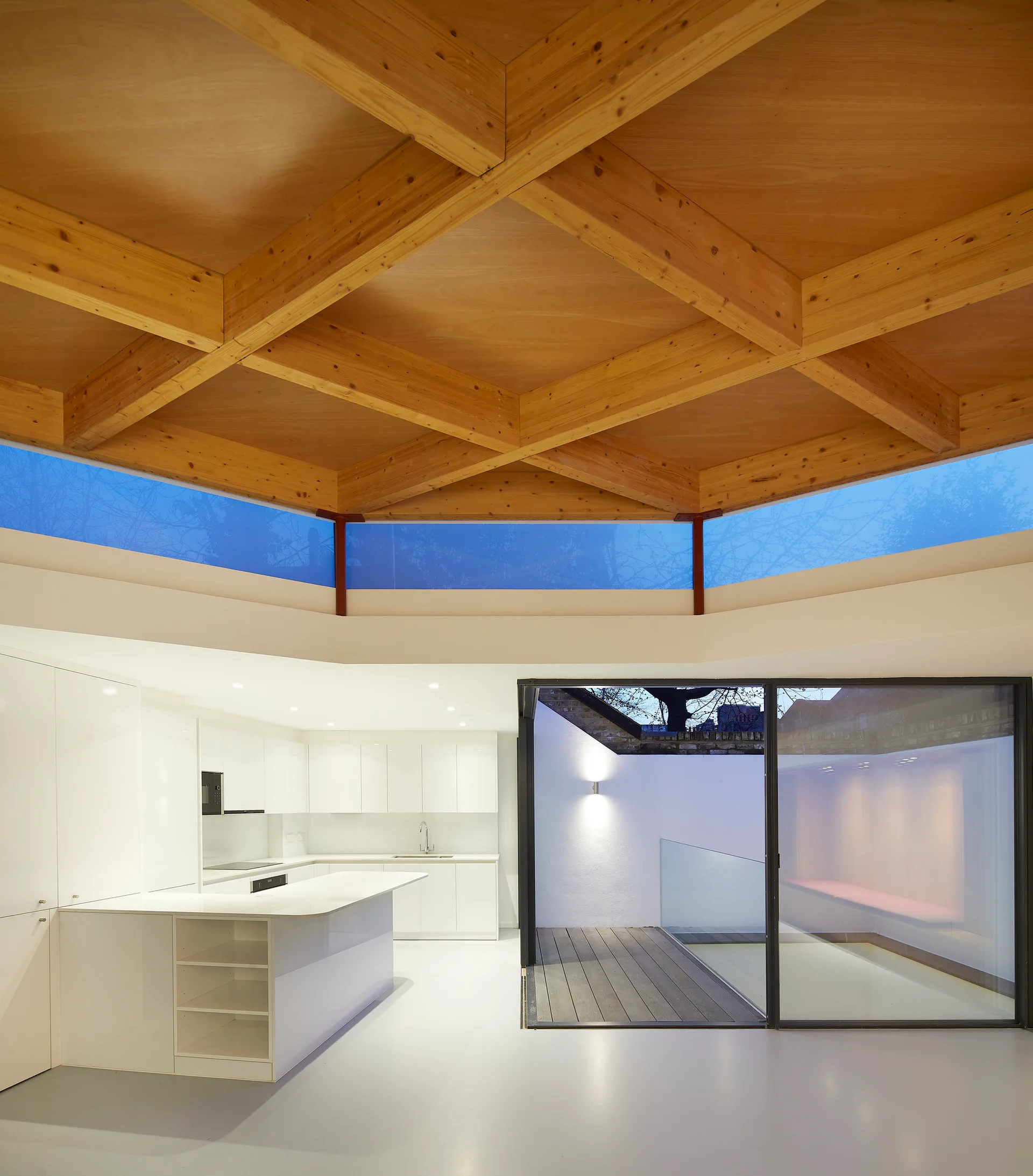 A modern white kitchen features a geometric wood joist ceiling, clerestory windows, sleek cabinets, and a sliding glass door opening to a courtyard.