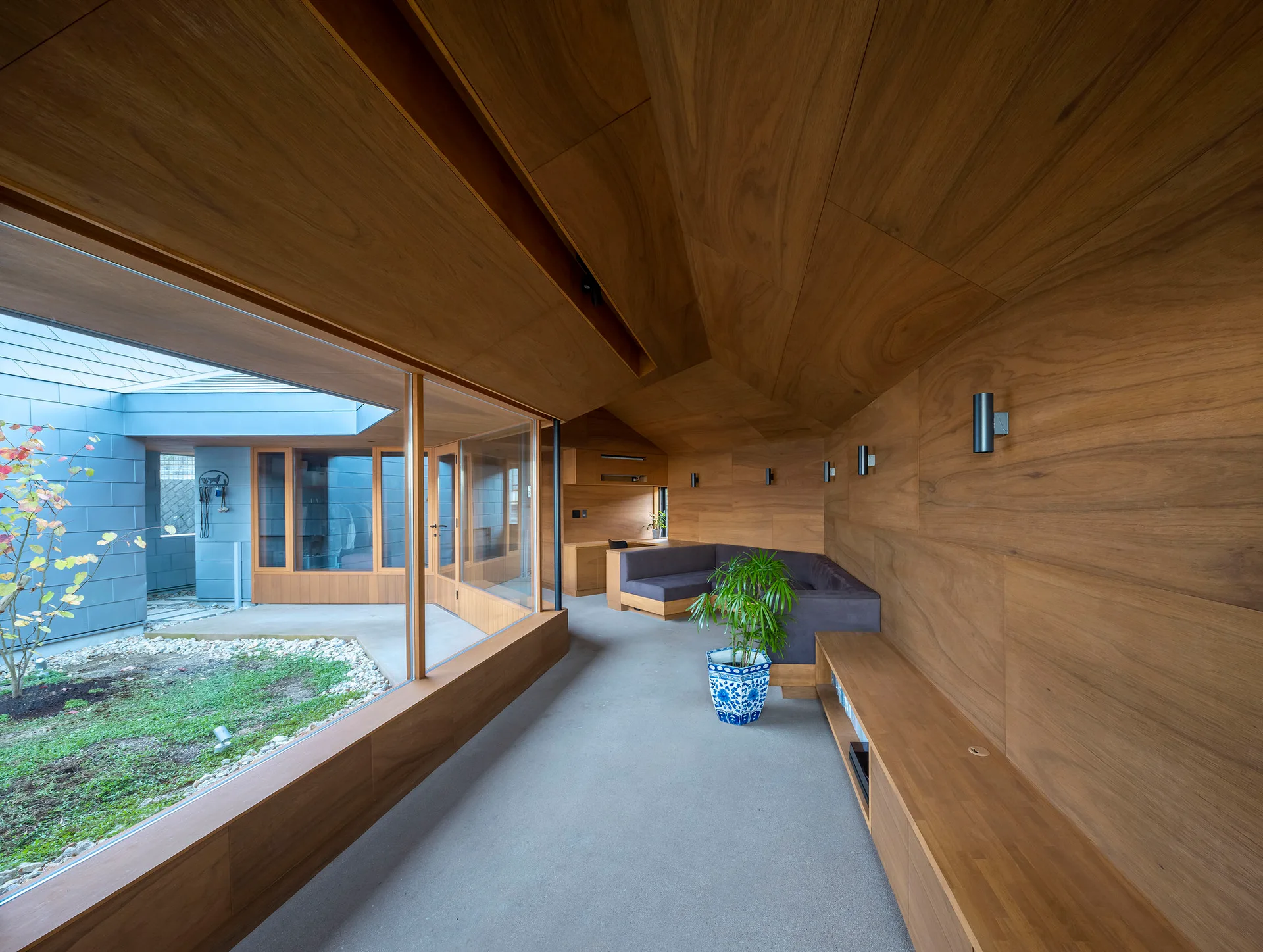 A minimalist room with extensive wood paneled walls and ceiling, a built-in sofa, and a large glass window facing an outdoor courtyard.