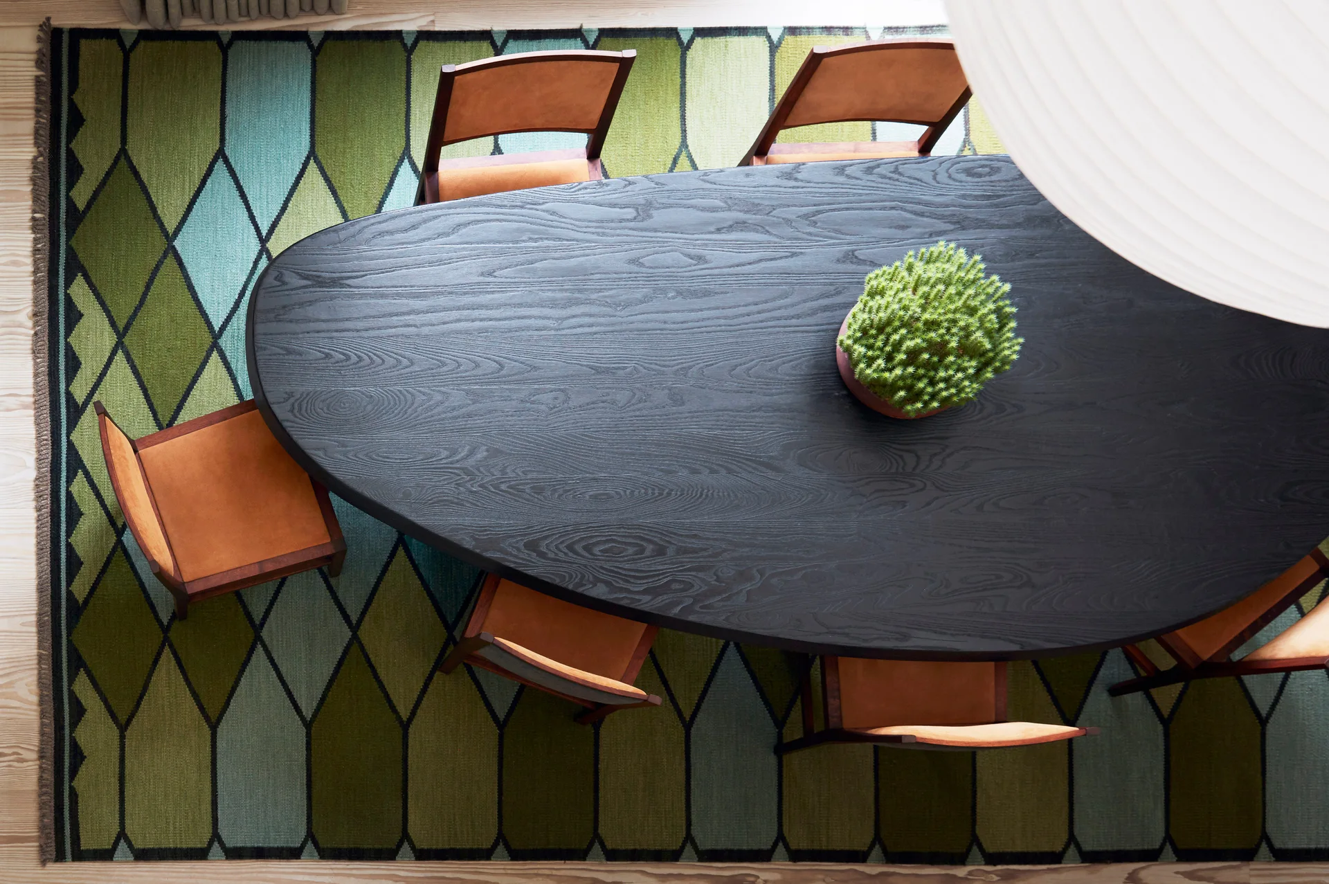 An overhead view of a matte black oval dining table and wooden chairs with tan seats on a green geometric patterned area rug.