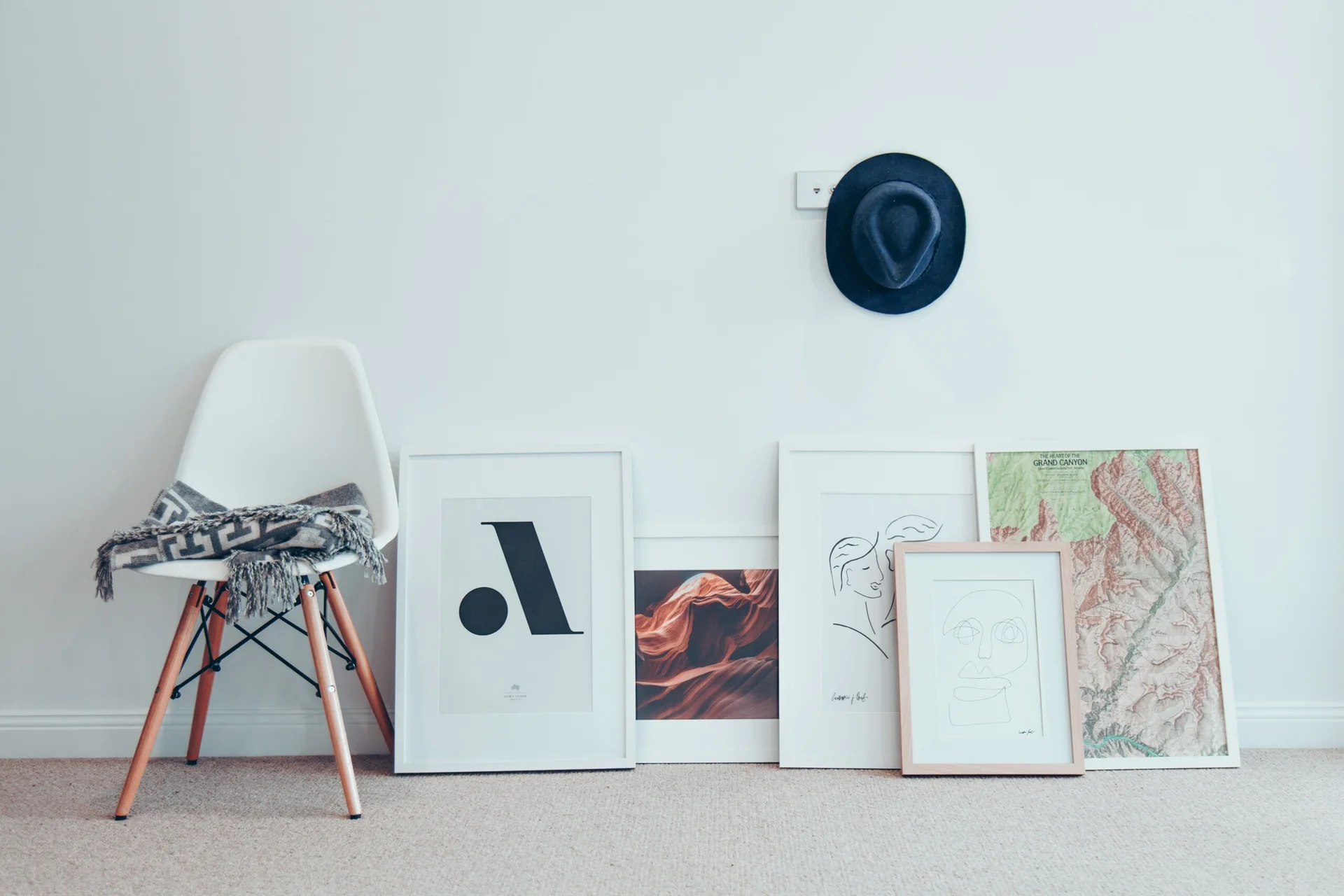 A white molded plastic chair draped with a gray blanket sits next to framed art prints leaning against a white wall below a hanging hat.