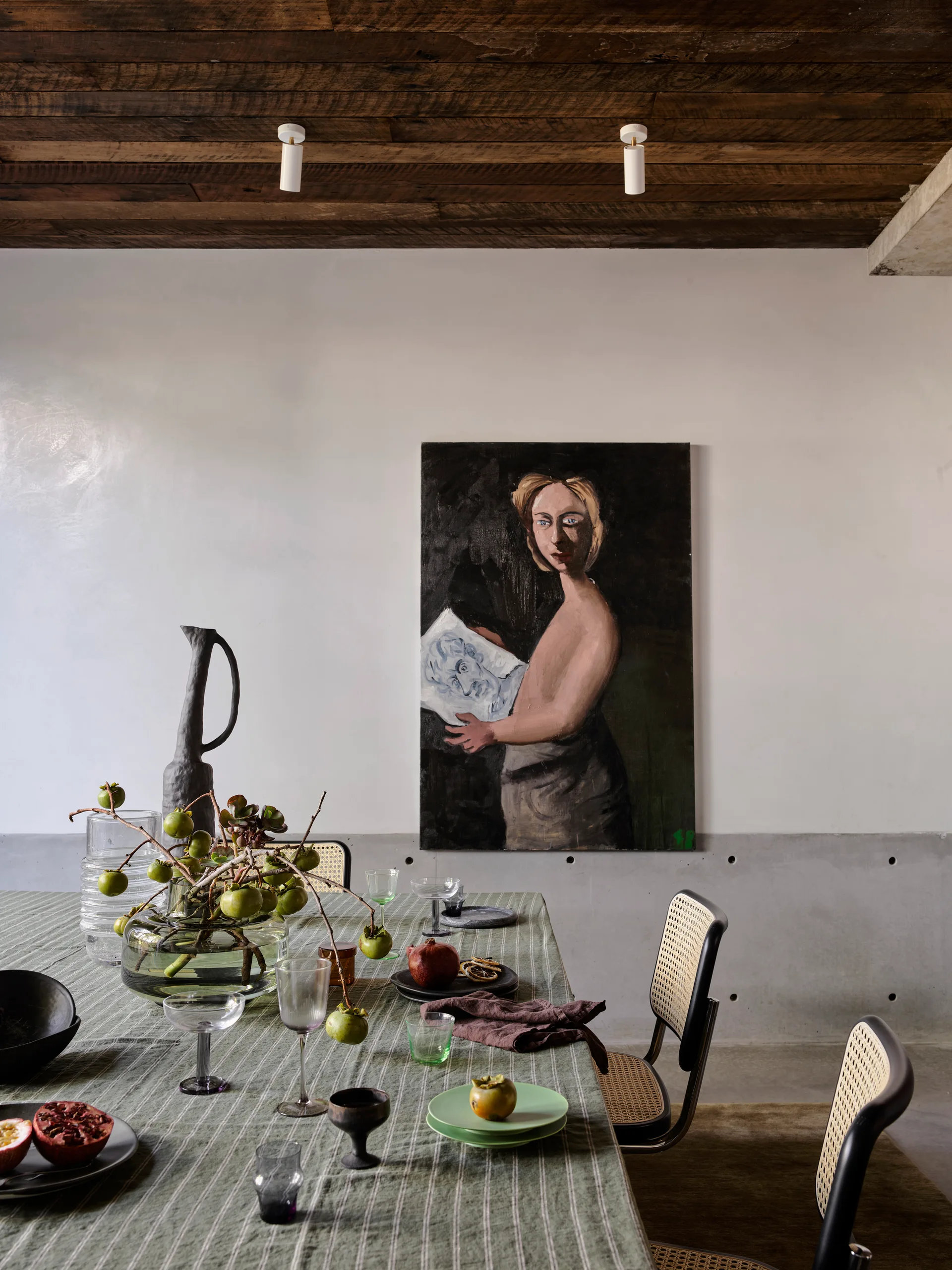 A dining table set with a striped tablecloth and fruit sits near cane-back chairs, a concrete wall, and a large portrait painting.
