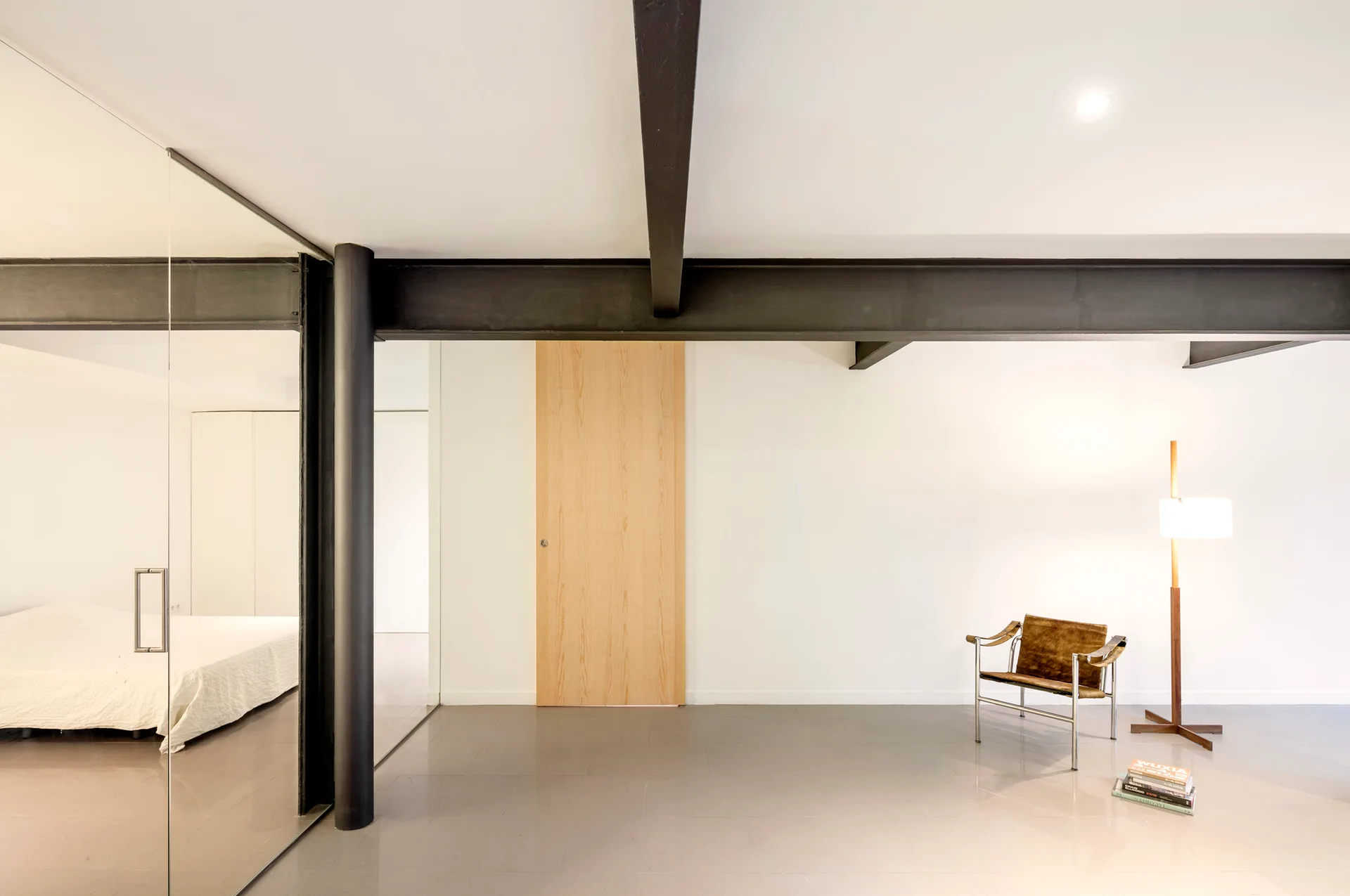An open interior with black steel beams, a glass partition enclosing a bedroom, a light wood door, and a brown leather armchair on grey tiled flooring.