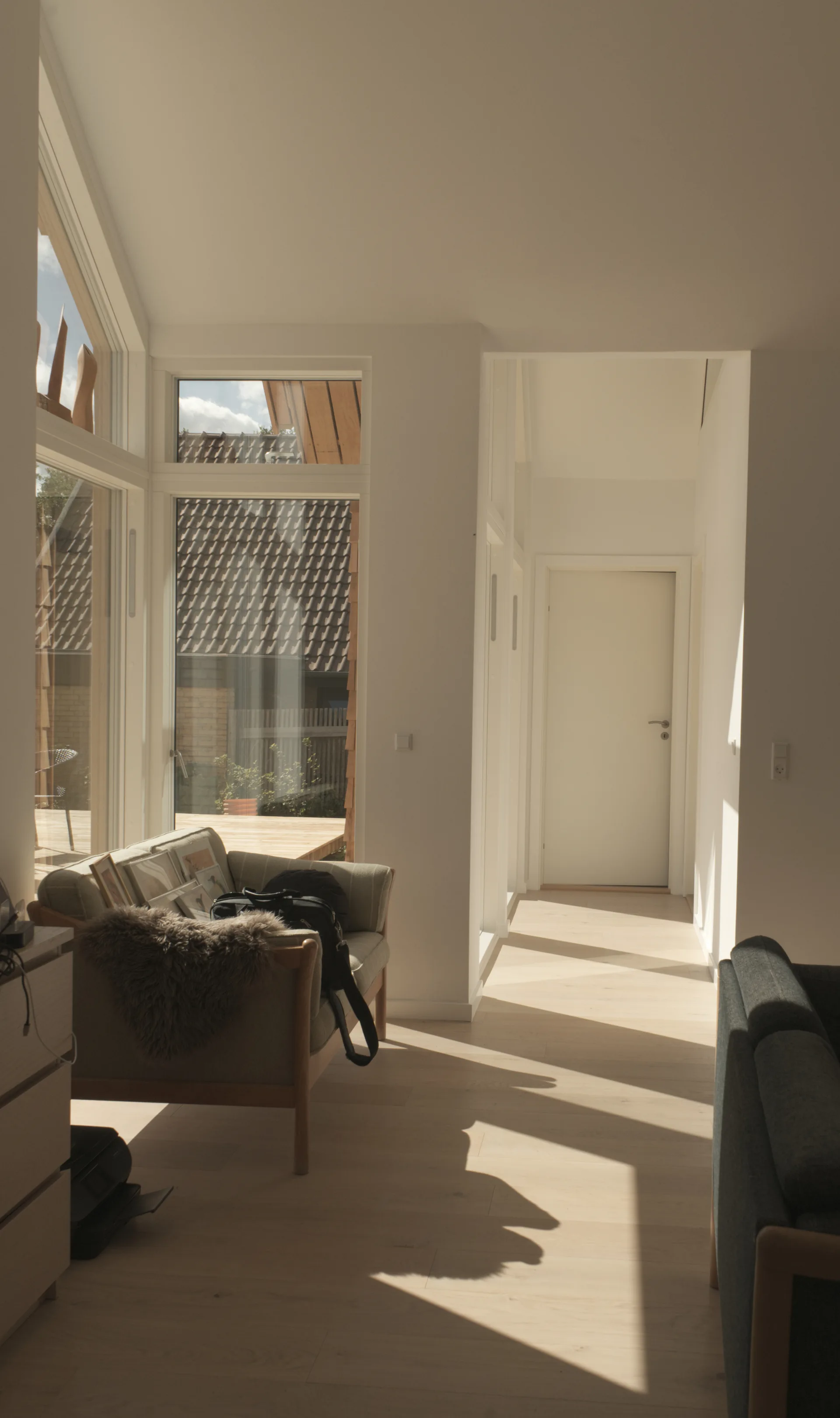 A bright Scandinavian living room featuring a neutral sofa, light wood flooring, and large windows that cast shadows.