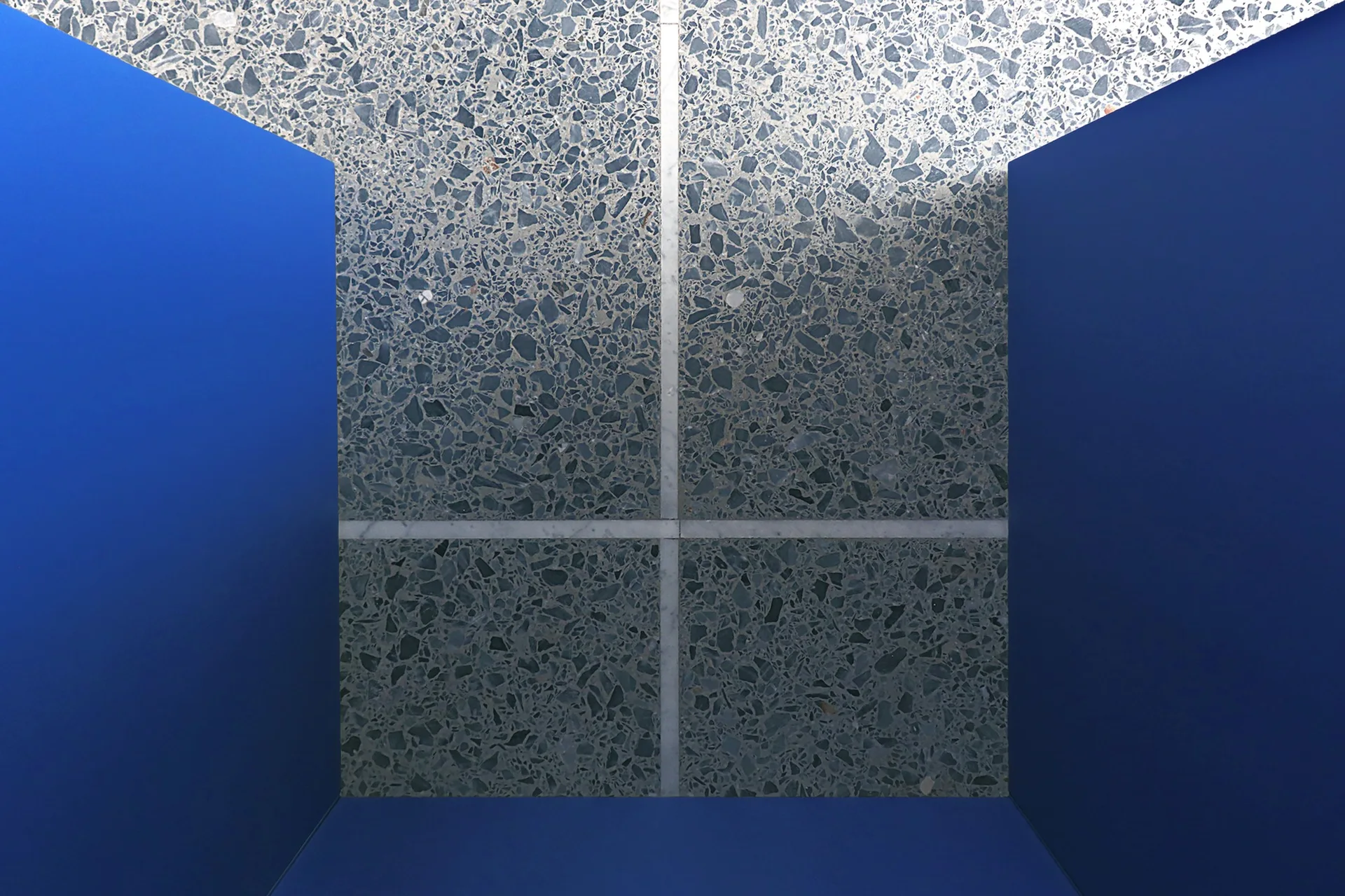 An interior corner features bold matte blue walls framing a speckled grey terrazzo floor divided by thin white lines.