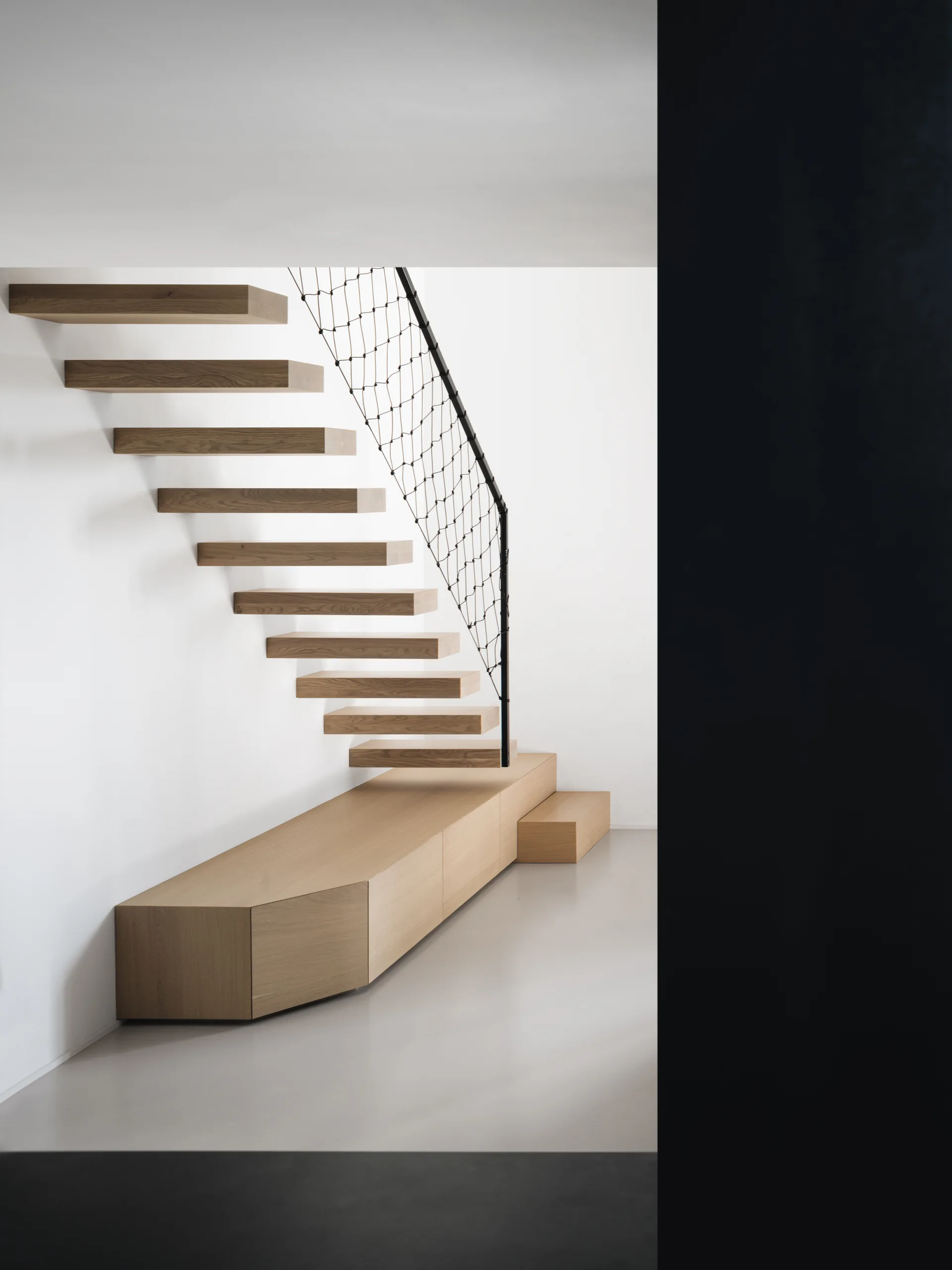 Minimalist floating wooden stairs rise above a low oak cabinet against a white wall with a black mesh safety netting railing.