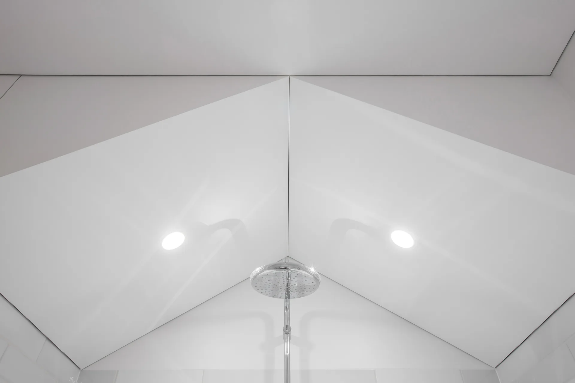 A chrome rainfall shower head sits below a white, sloped geometric ceiling with integrated recessed lighting in a modern bathroom.