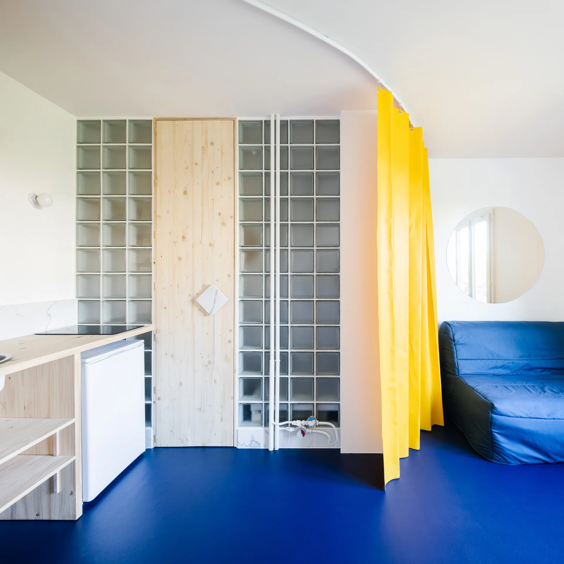 A compact studio interior featuring a vibrant blue floor, a yellow curtain on a curved ceiling track, and a glass block wall surrounding a wood door.