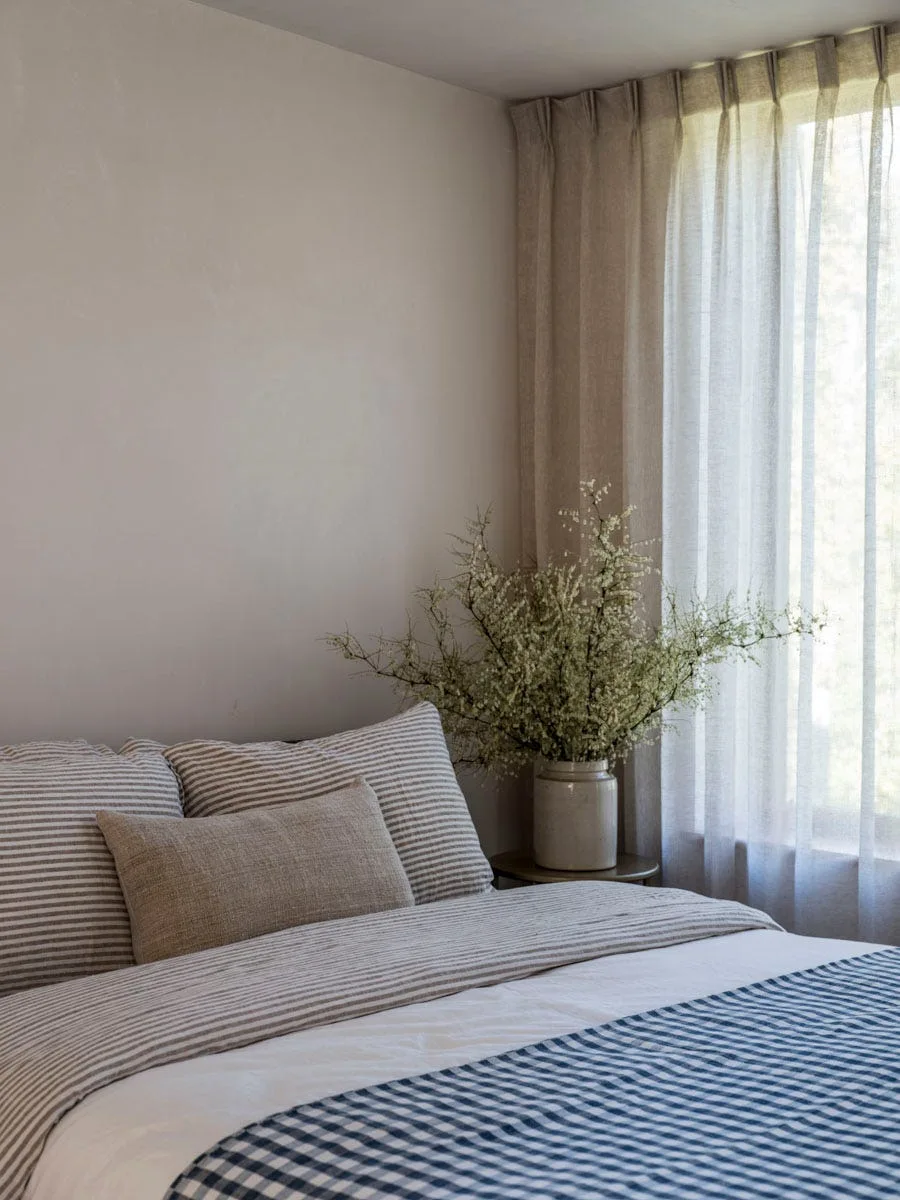 A bed with striped pillows and a blue checkered blanket sits beside a window draped in sheer beige curtains with a vase of branches on the nightstand.