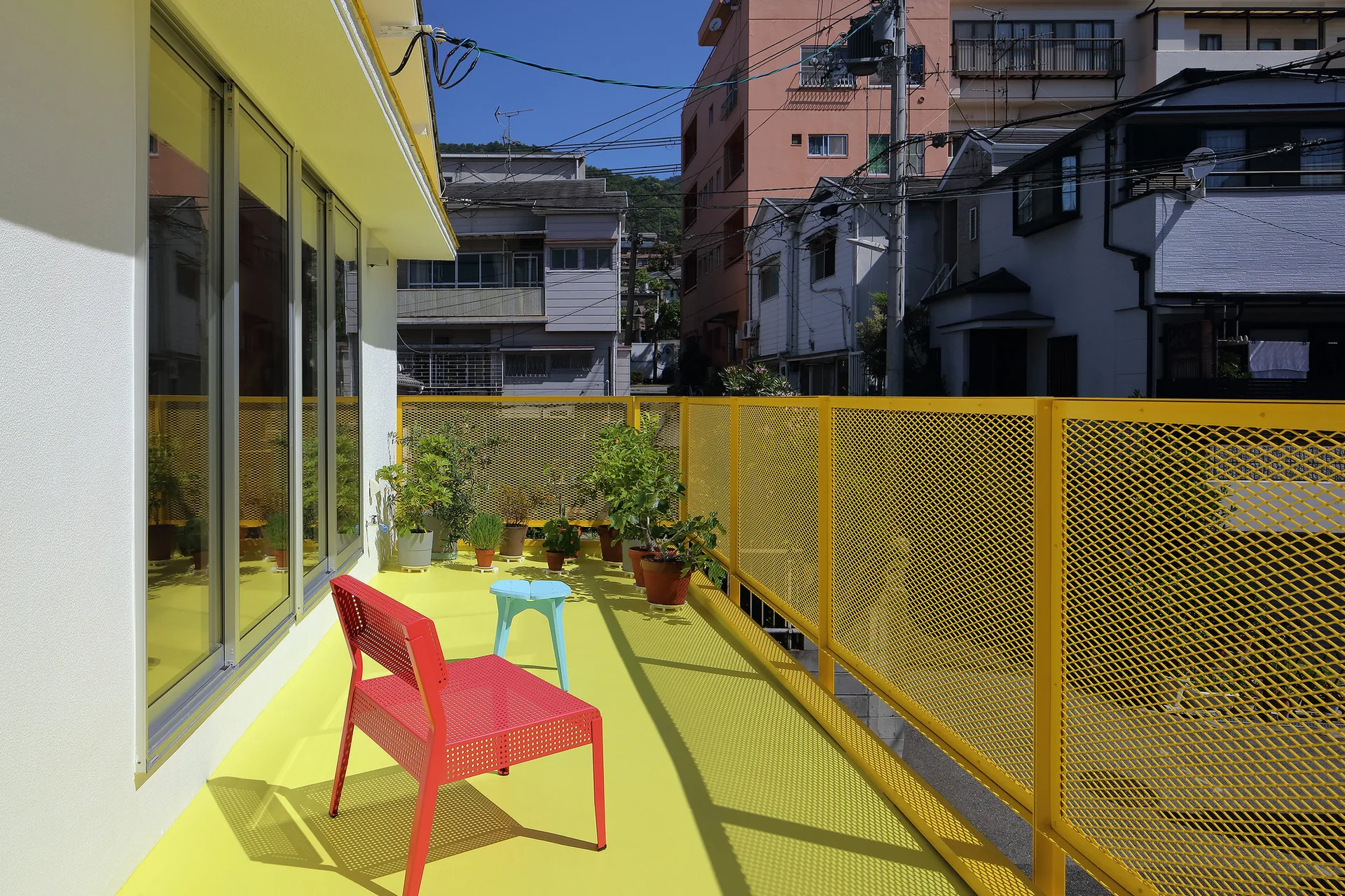 A bright yellow outdoor balcony features a red perforated metal chair, a light blue stool, and green potted plants next to a yellow mesh railing.
