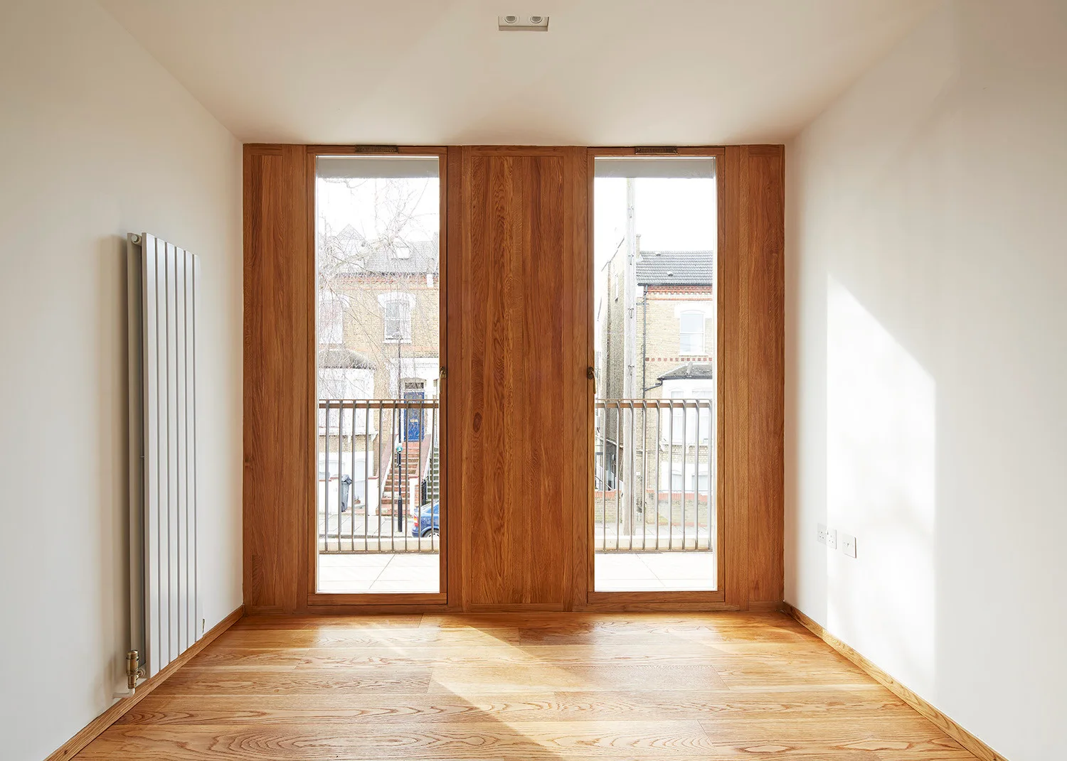 An empty minimalist room features natural wood floors, plain white walls, a slim white vertical radiator, and tall wood-framed windows.