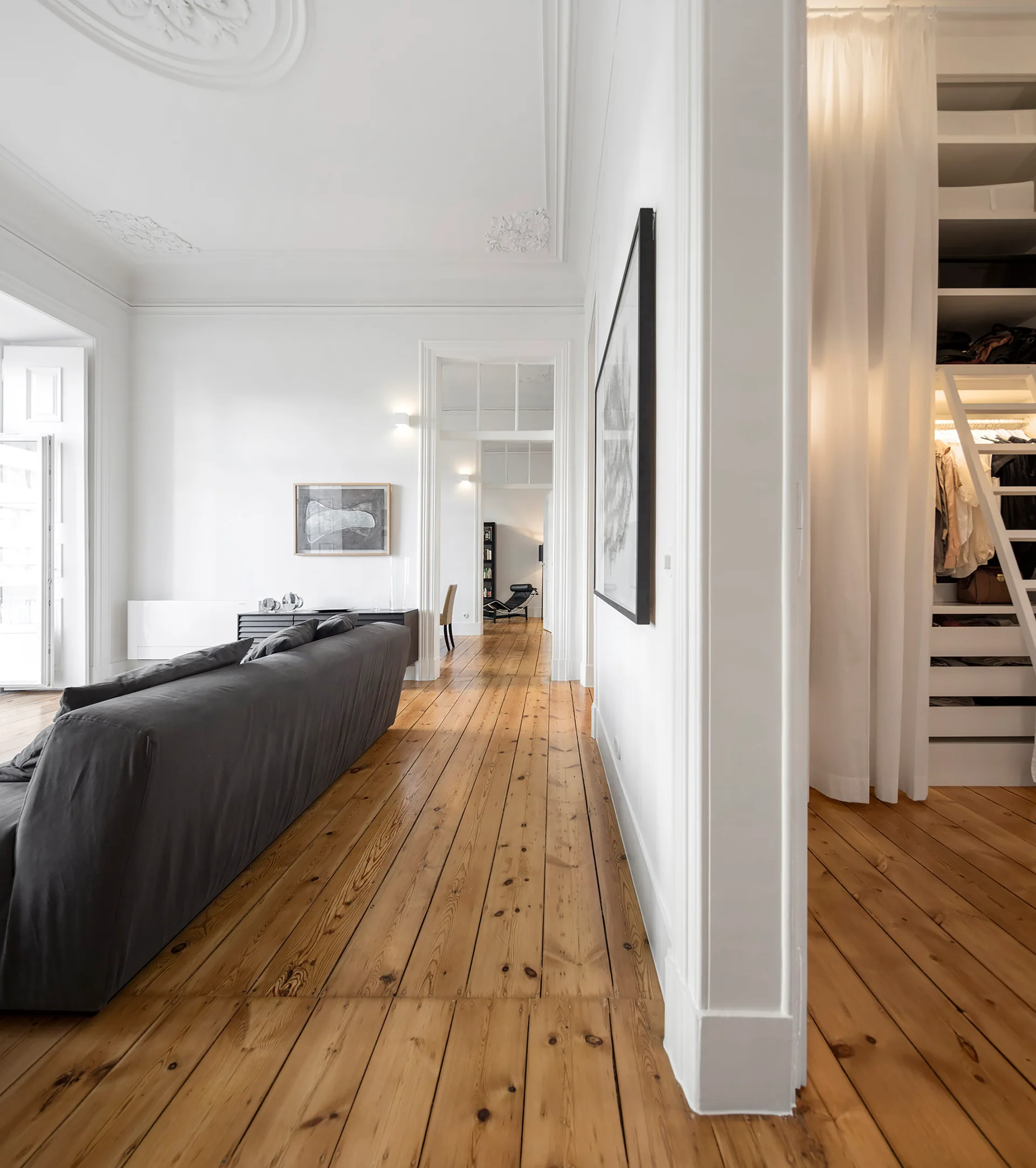 A bright white minimalist living room with wide-plank pine floors, a grey sofa, and a closet screened by a white curtain.