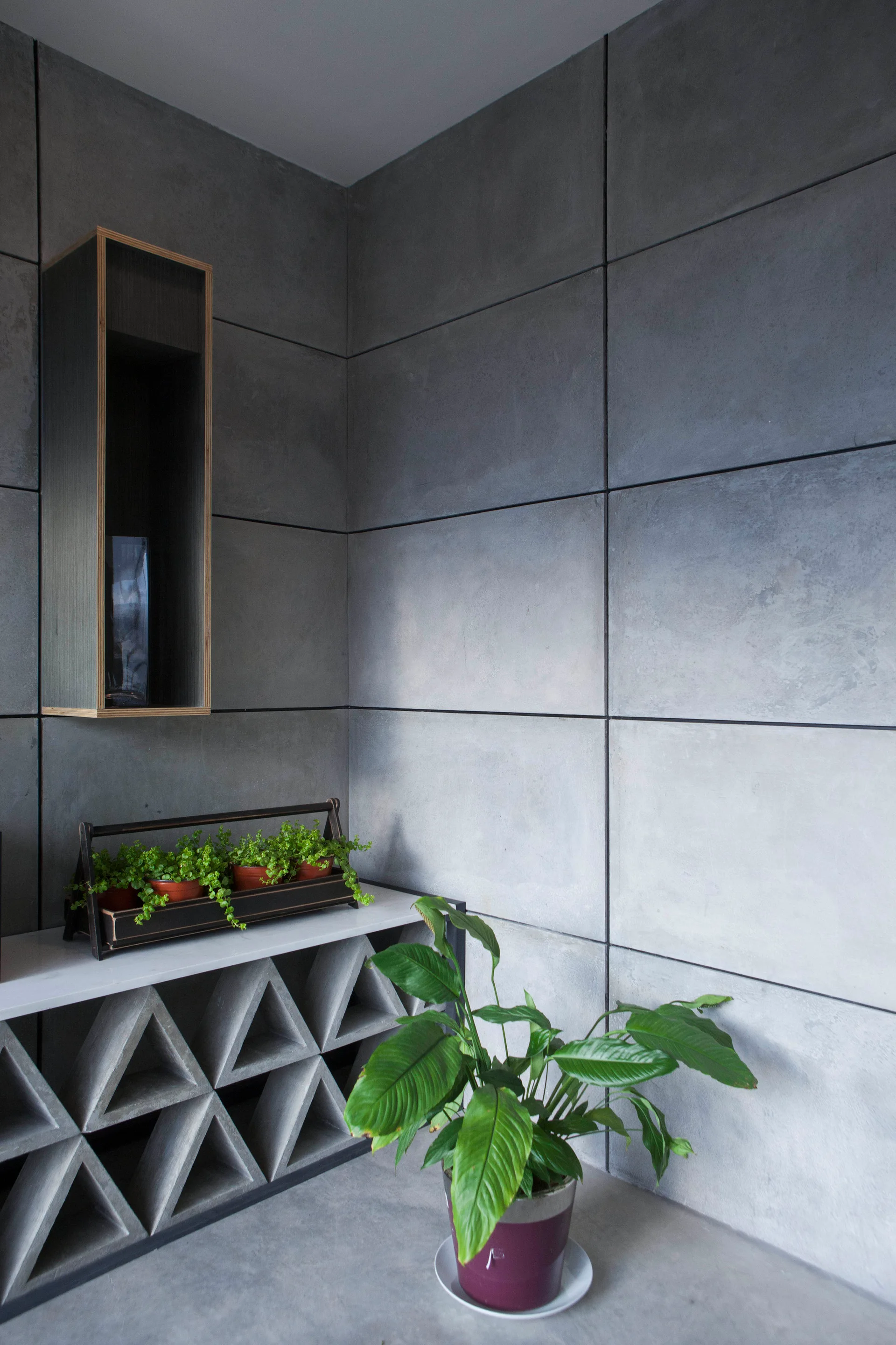 A modern indoor corner features modular grey concrete wall panels, a console table with a geometric triangular base, and several green potted plants.