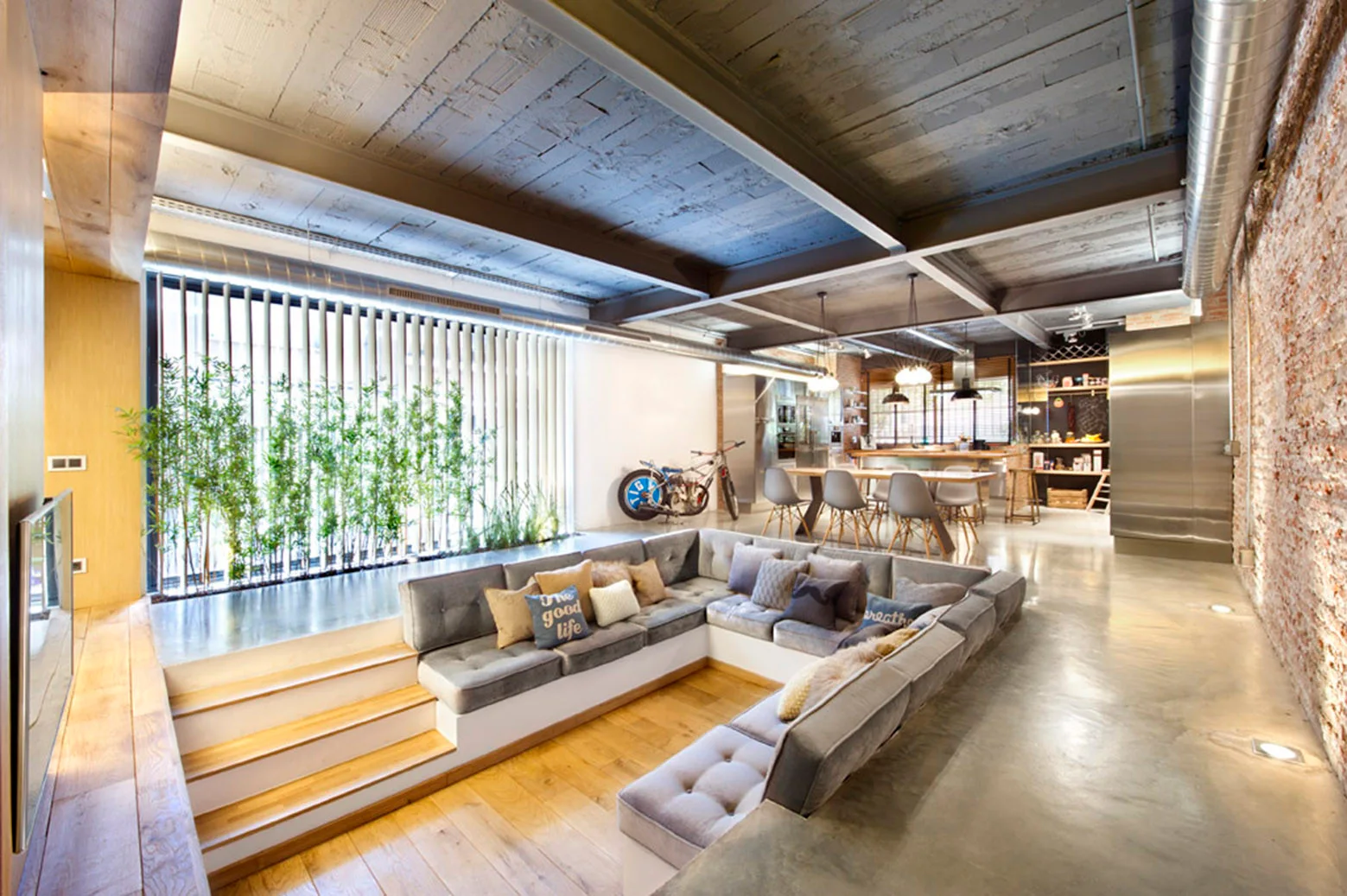 A spacious industrial loft features a sunken conversation pit with a grey sectional sofa, wood steps, and polished concrete floors.
