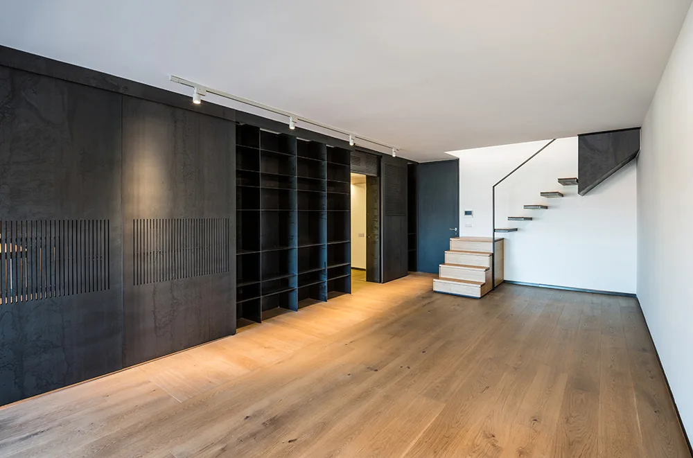 A minimalist interior featuring a large black built-in shelving unit with sliding slatted panels, wood flooring, and a floating wood staircase.