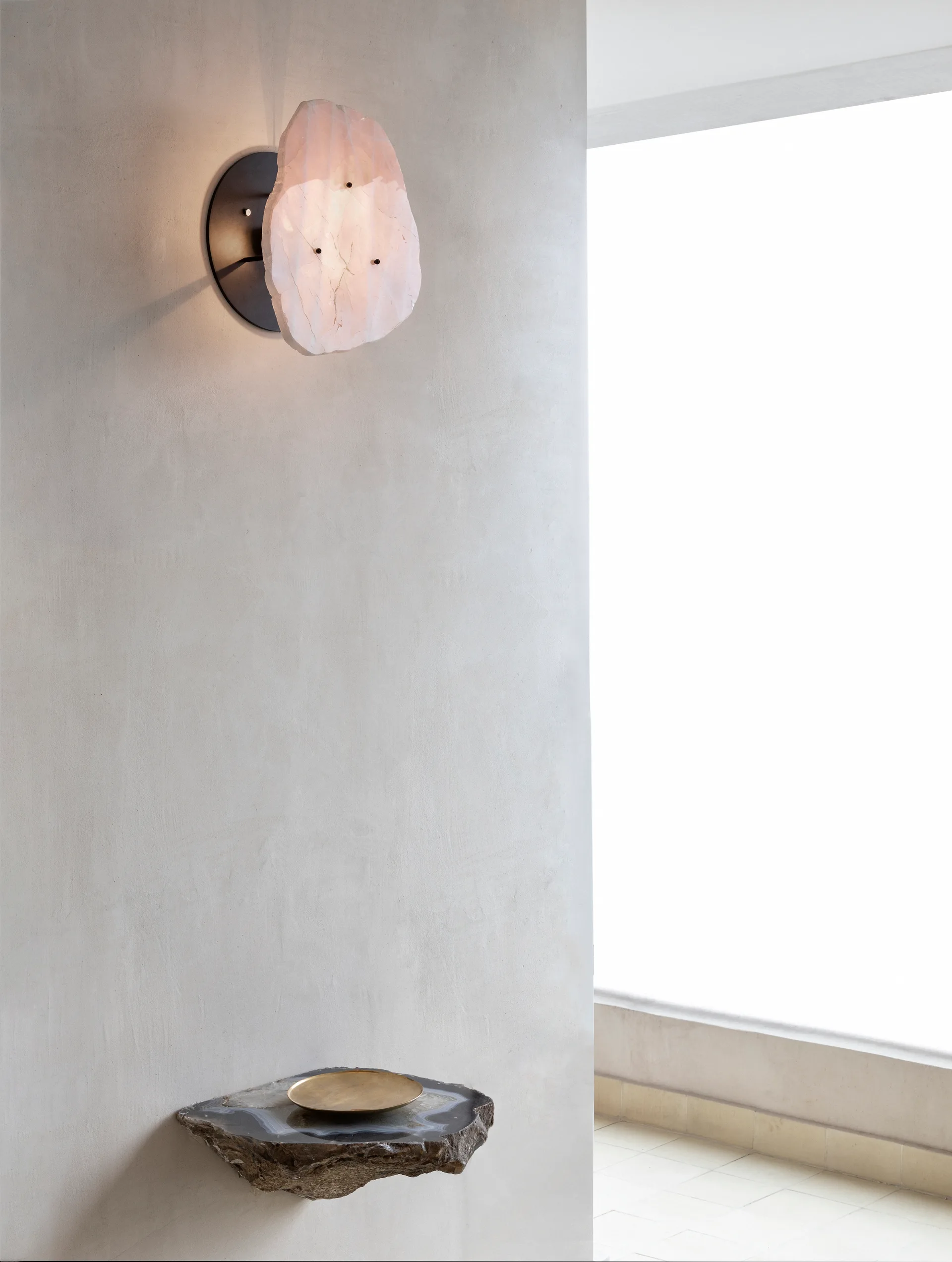 A backlit raw quartz wall sconce glows above a small stone floating shelf with a gold dish on a smooth plaster wall.