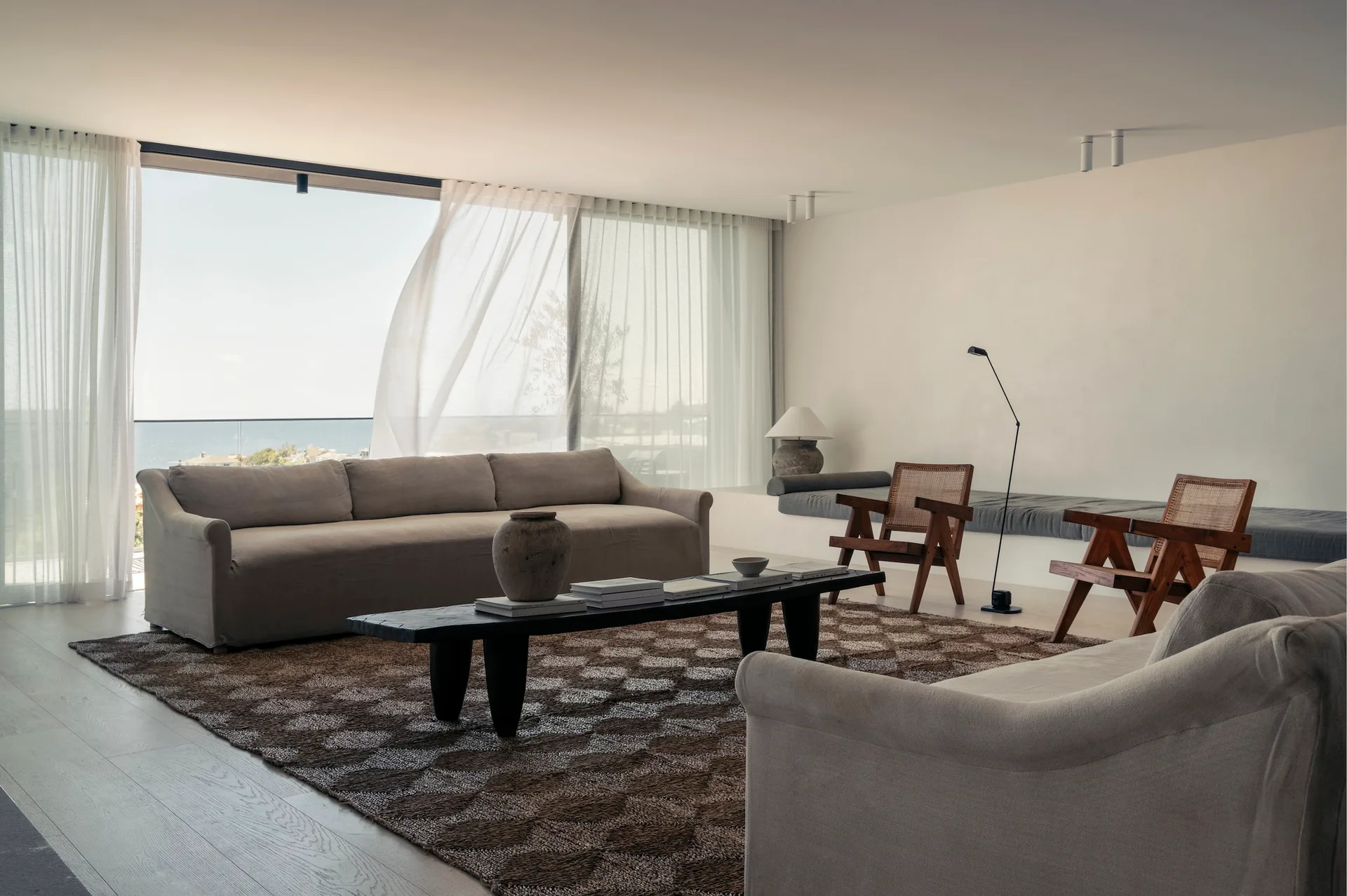 Beige sofas and cane armchairs surround a dark wood coffee table on a geometric rug in a bright room with large windows and sheer curtains.