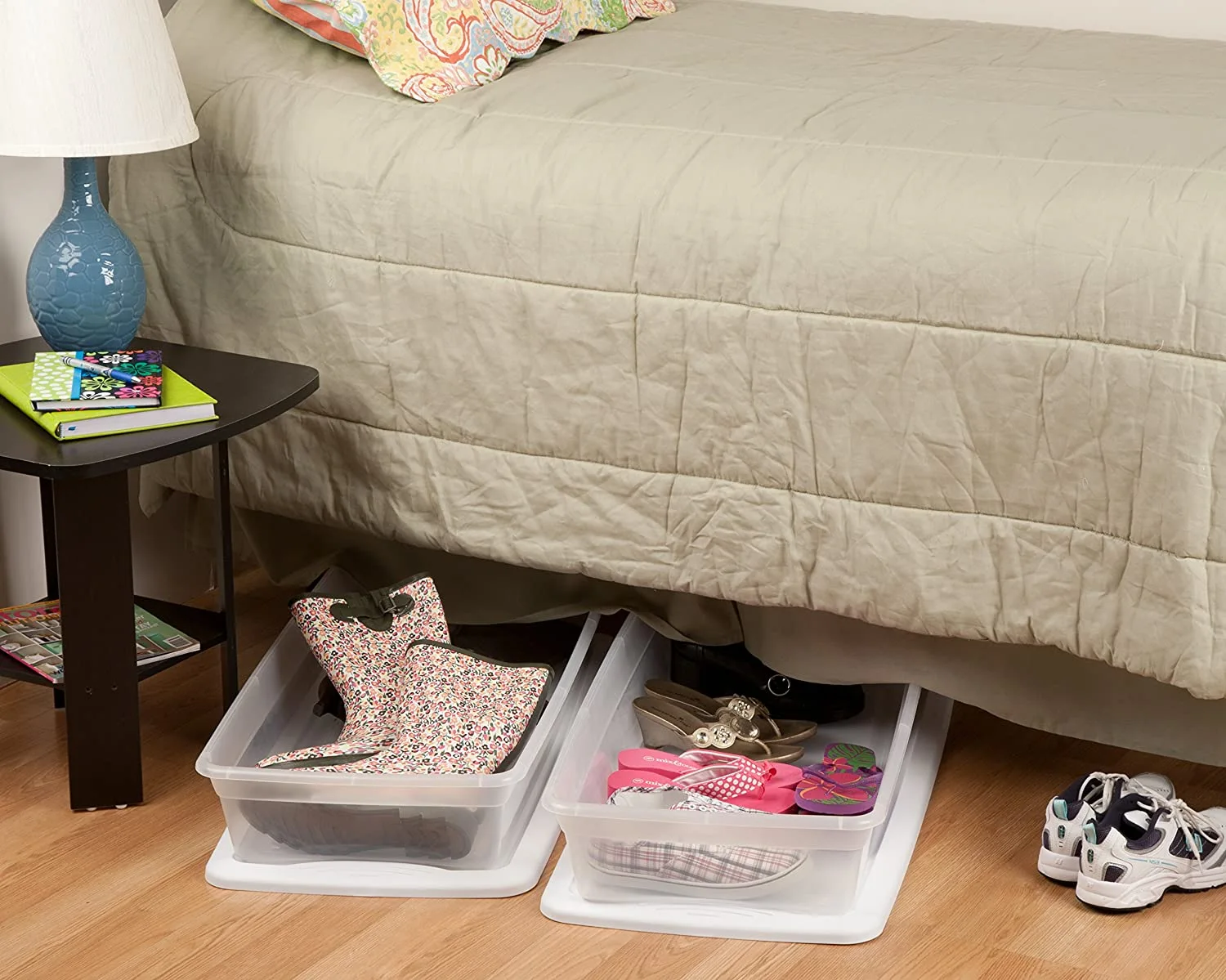 Two translucent plastic storage bins filled with shoes slide out from under a bed with a beige quilted coverlet next to a dark wood nightstand.