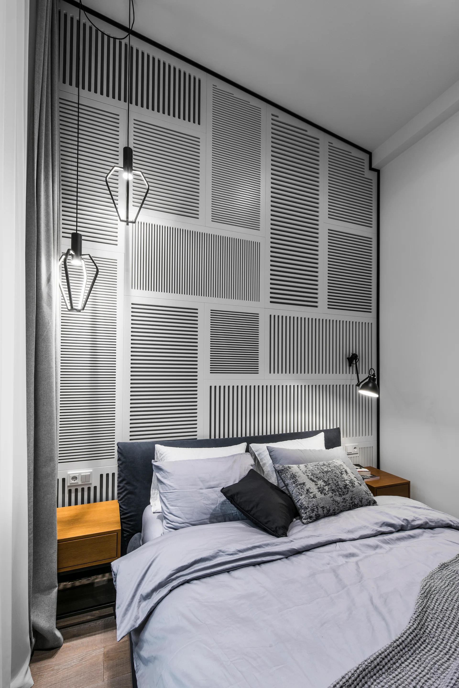A bed with grey linens sits against a textured white slatted accent wall, flanked by wooden nightstands and hanging geometric pendant lights.