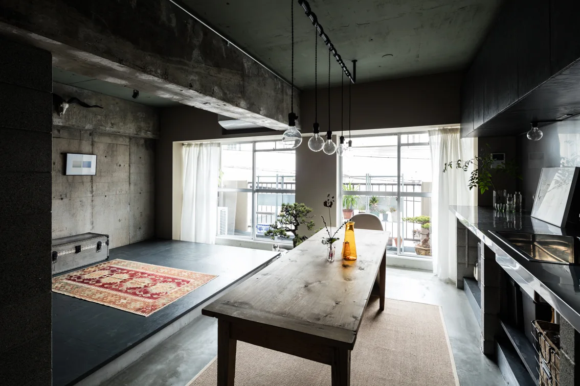 An industrial dining space features a raw wooden table, exposed concrete walls, a raised platform with an oriental rug, and sheer curtains.