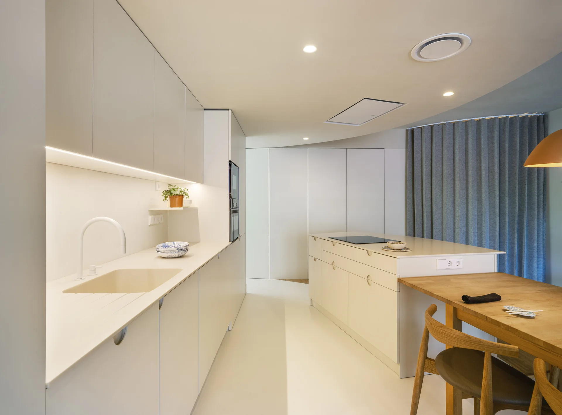 A minimalist off-white kitchen features seamless cabinetry, a central island with an integrated wooden dining table, and a grey curtain.