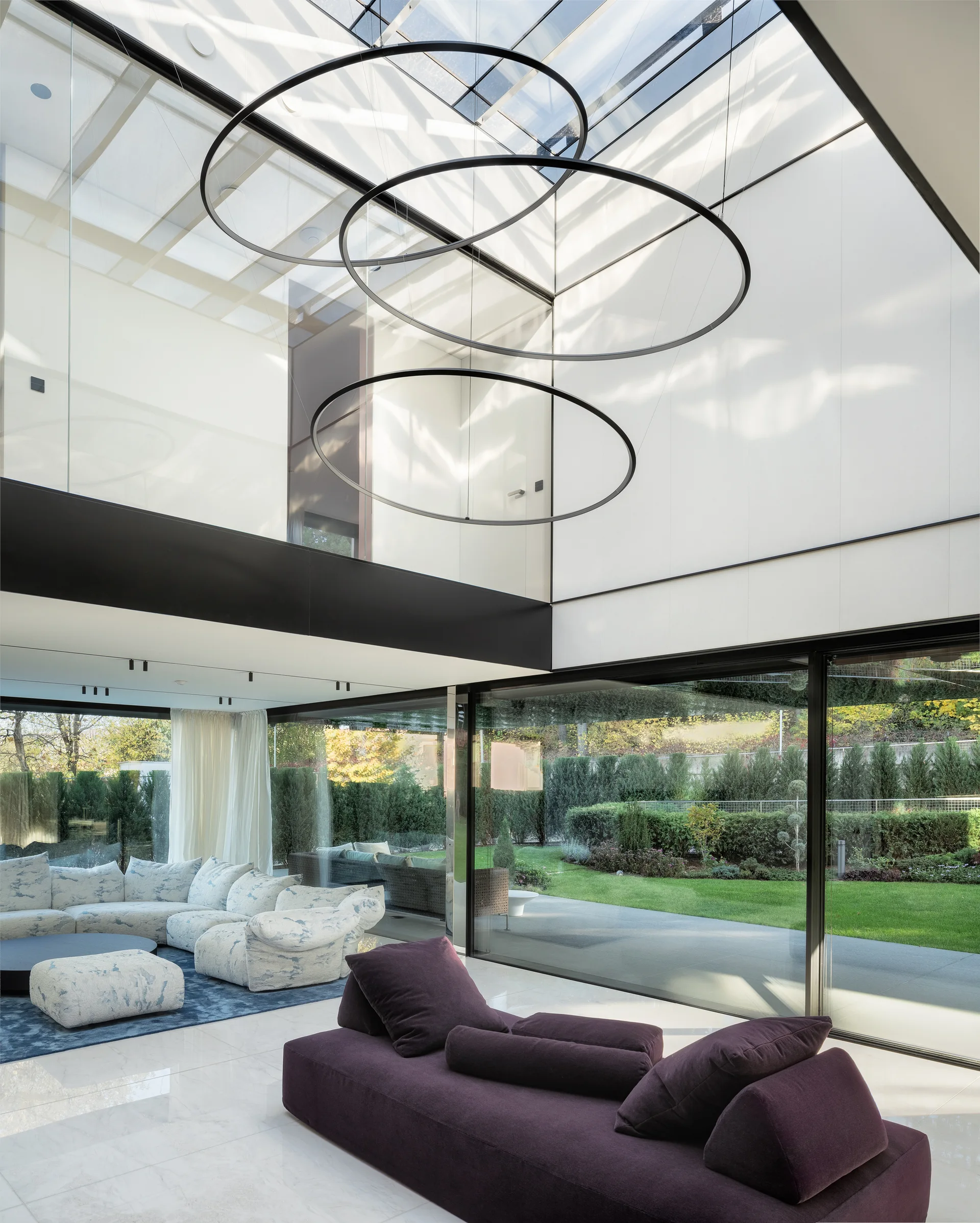 A modern double-height living room featuring floor-to-ceiling glass walls, a purple sofa, a patterned sectional, and large circular black pendant lights.