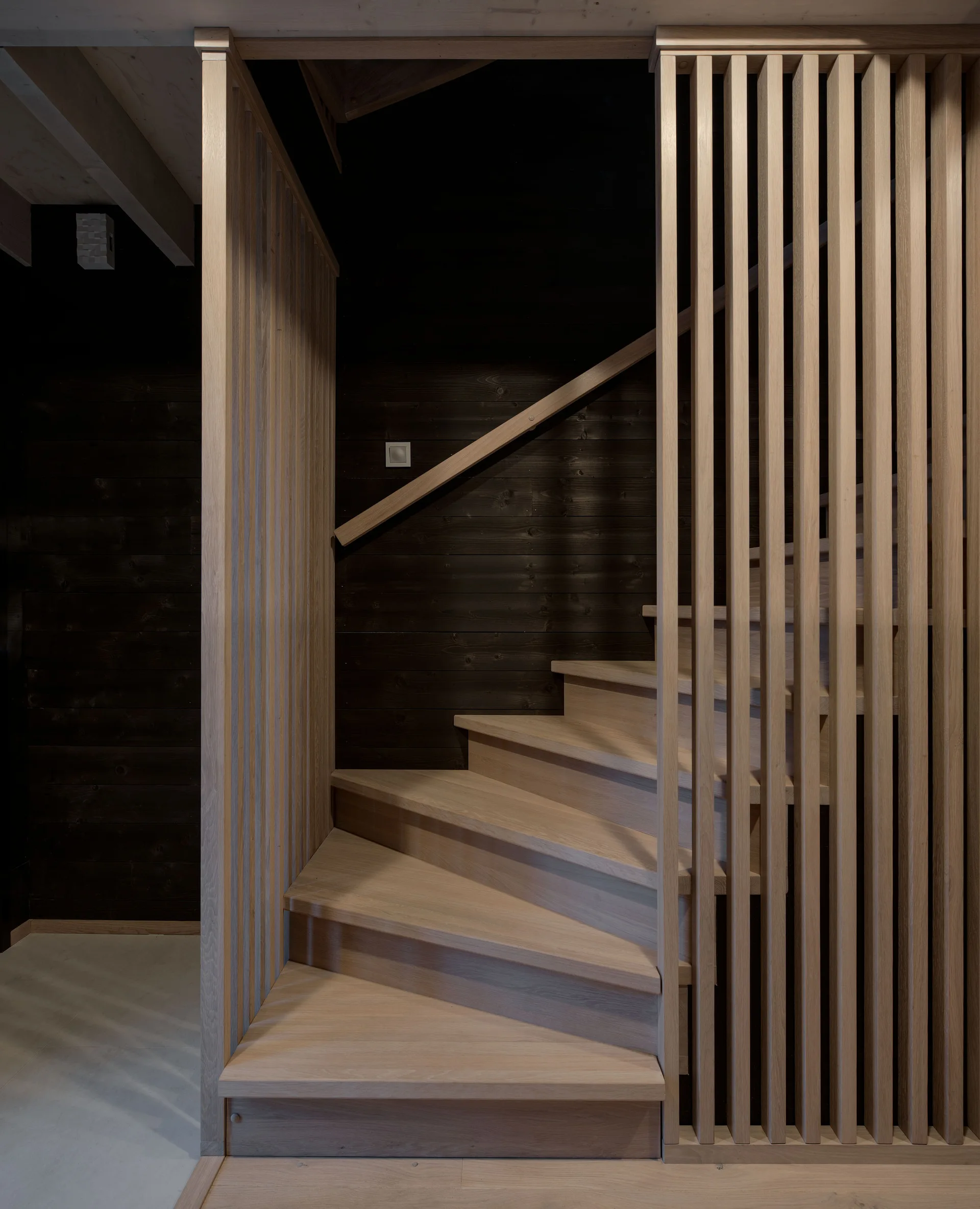 A light wood staircase with a vertical slatted timber screen is set against dark horizontal plank wall paneling.
