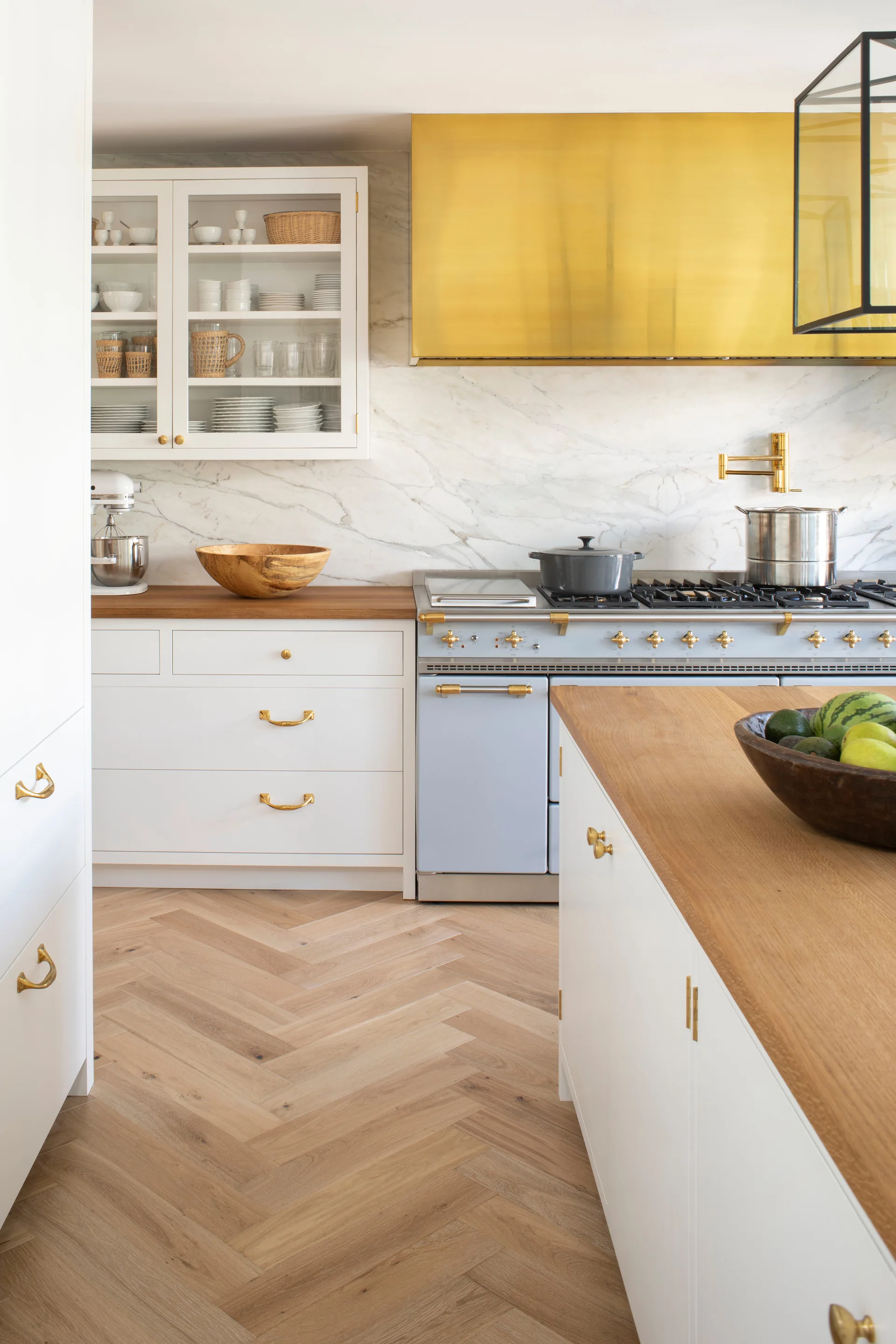 A bright kitchen features white cabinets with gold pulls, a blue stove under a gold range hood, marble backsplash, and herringbone wood flooring.
