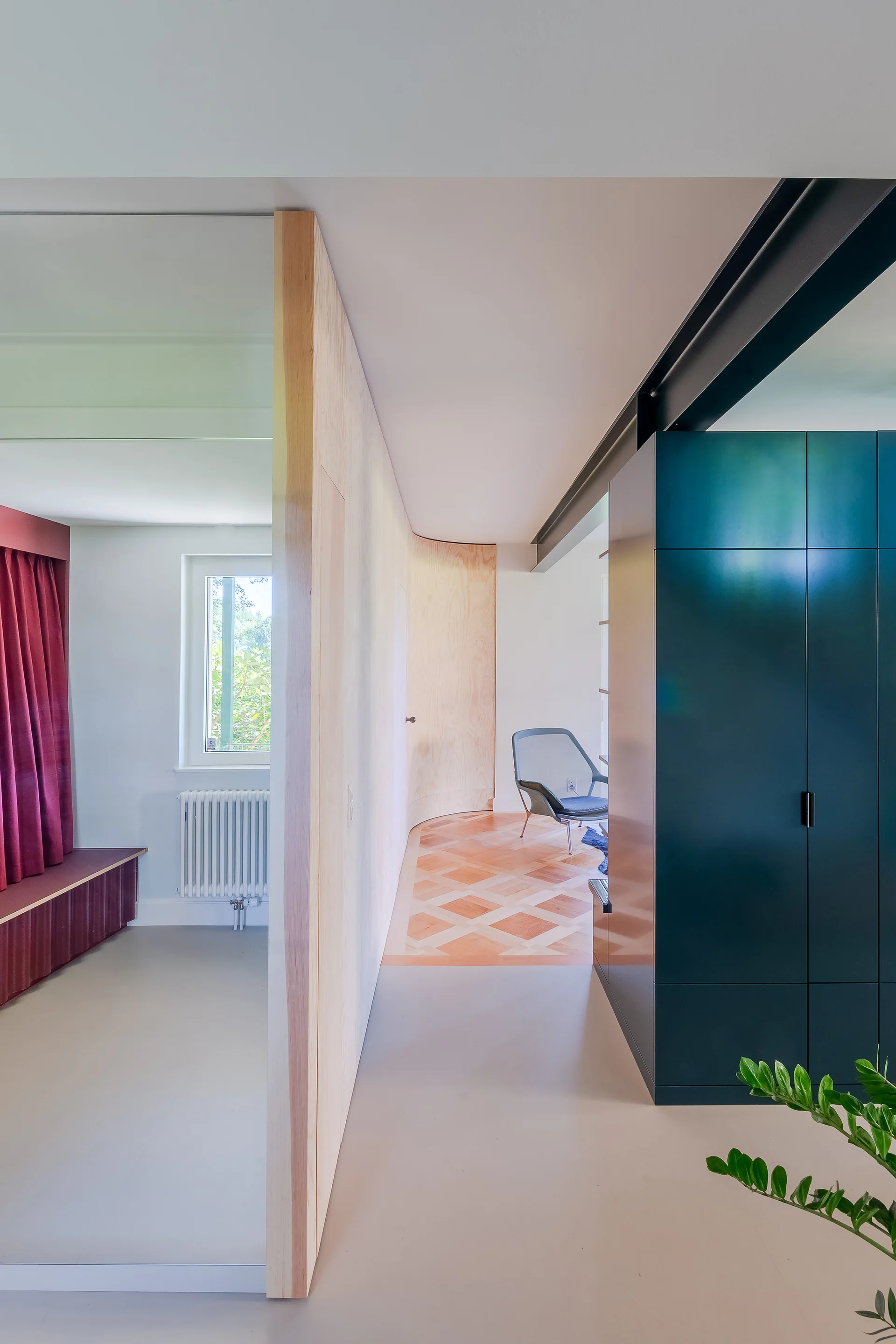 A modern hallway features a curved light wood partition wall, a large dark green storage cabinet, and a transition to geometric wood flooring.