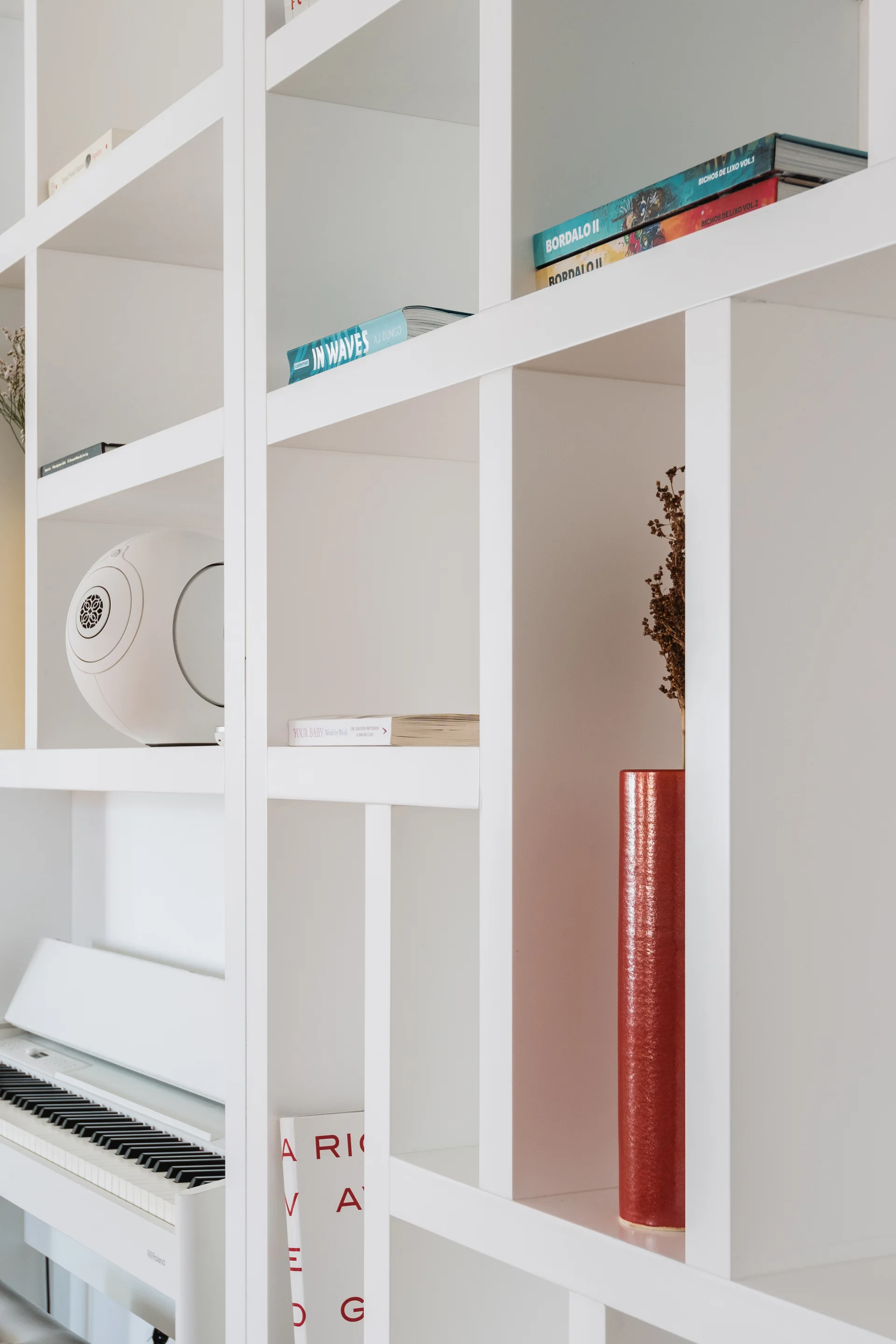 A matte white grid bookshelf displaying books, a round white speaker, and a tall red vase with a digital piano built into the lower section.